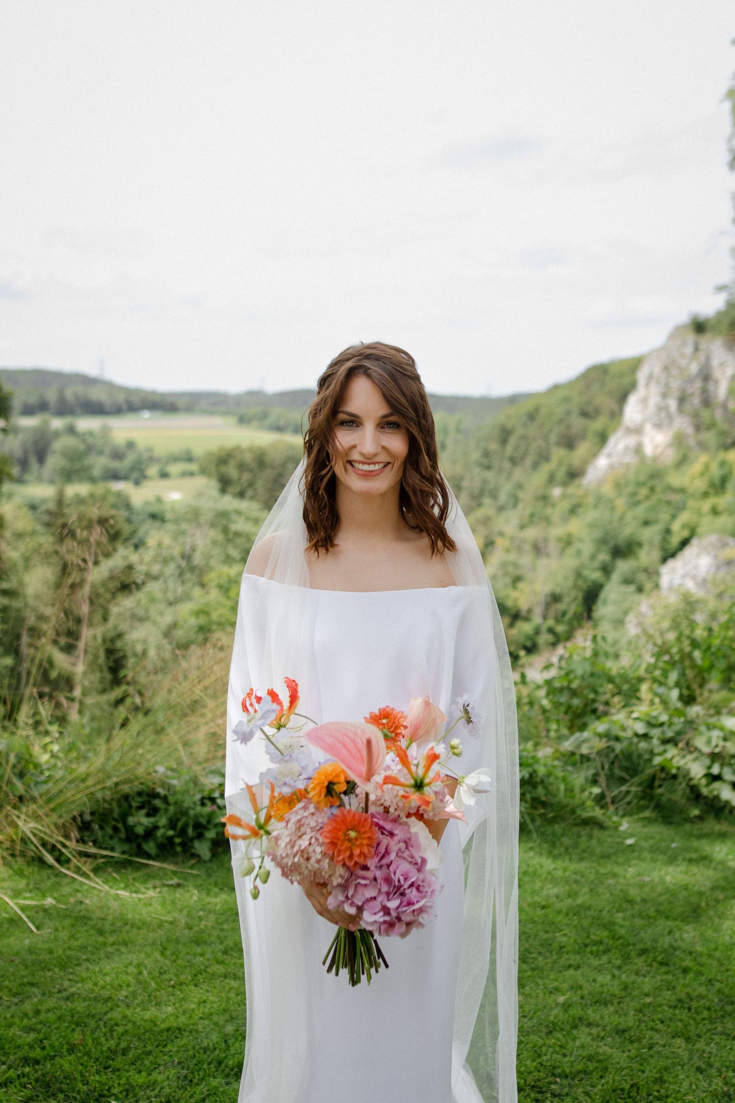 A bride in an off-shoulder gown holds a vibrant bouquet of coral and pink flowers against a scenic landscape.