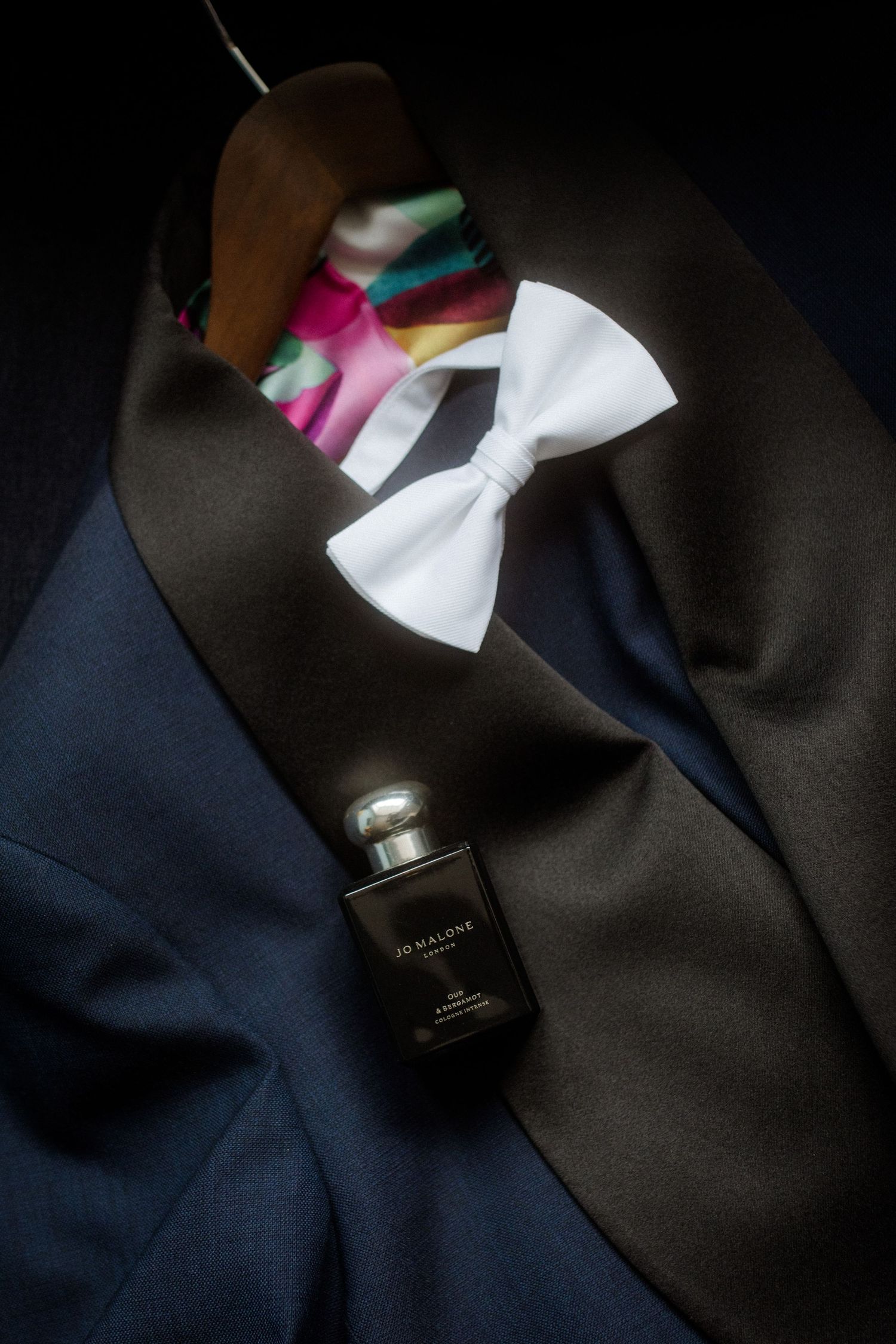 A formal suit with white bowtie and perfume bottle arranged on dark fabric.