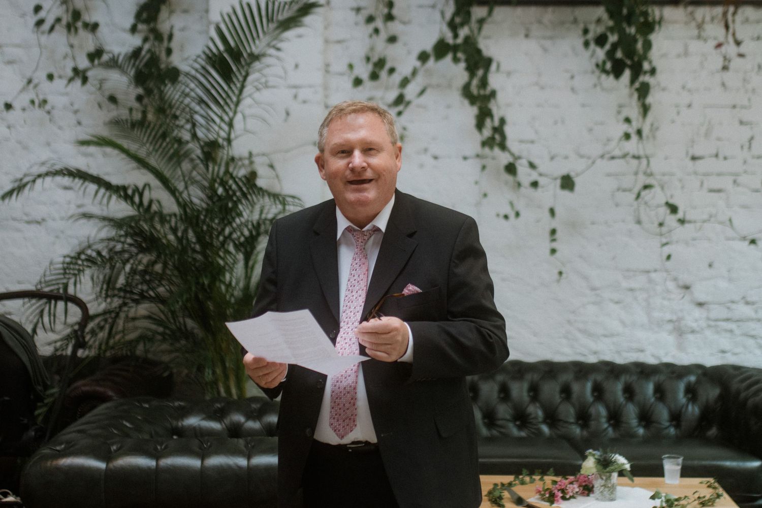 A gentleman in a dark suit reads from papers at an event with a white brick wall backdrop.