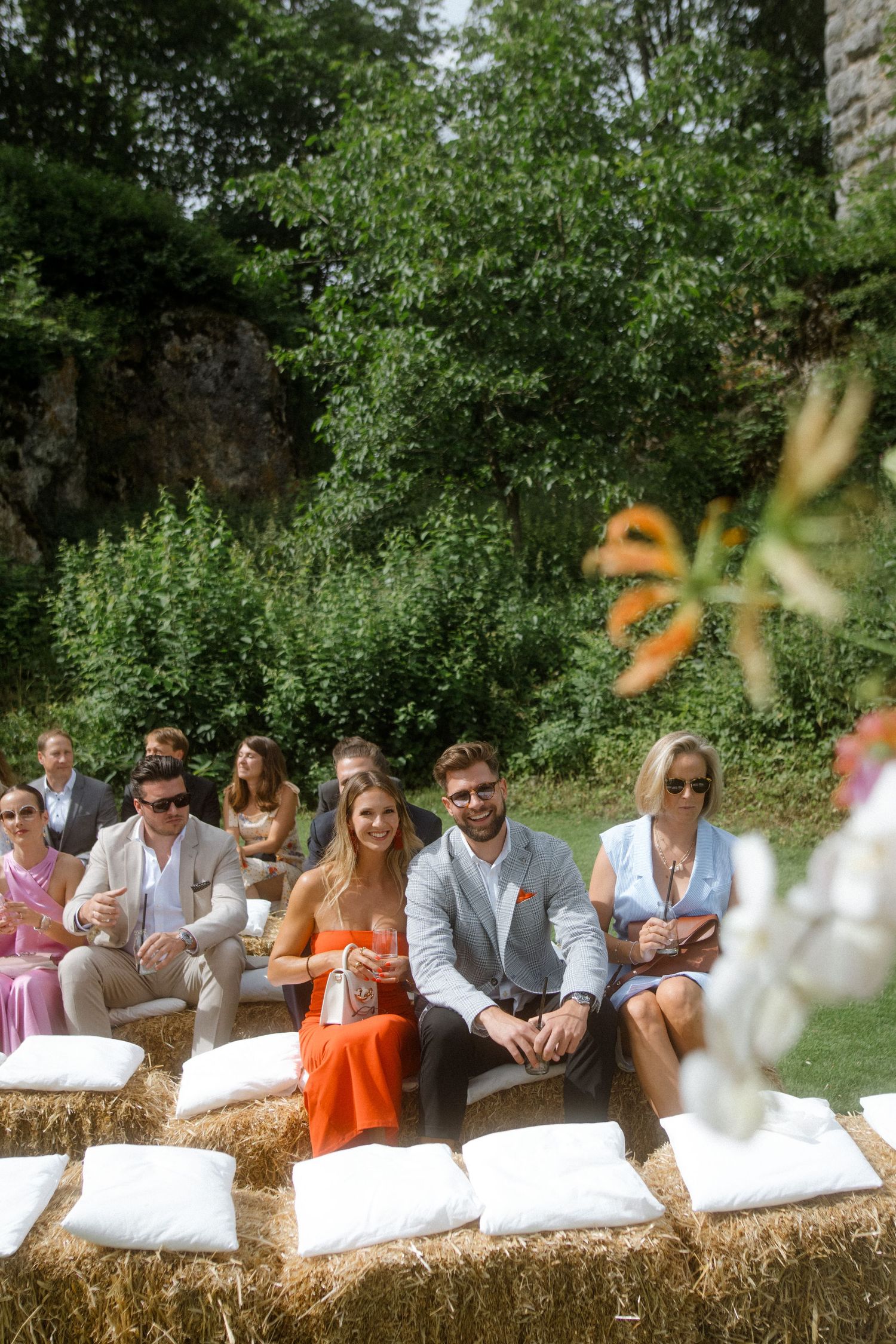 Wedding guests seated on hay bales decorated with white cushions in a garden setting.