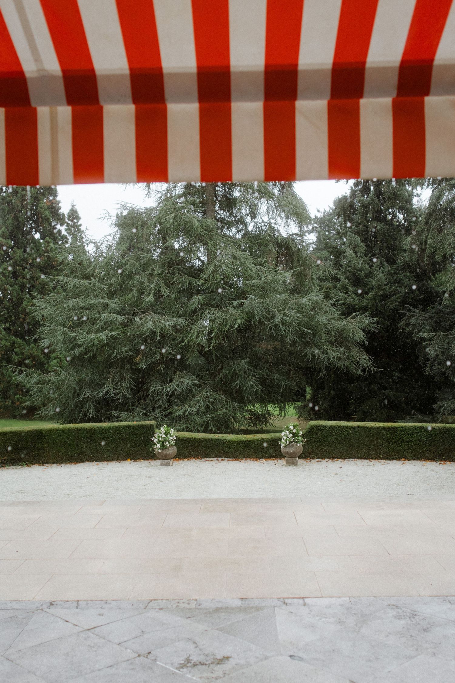 Red and white striped awning over a stone patio with manicured hedges and garden in the background.