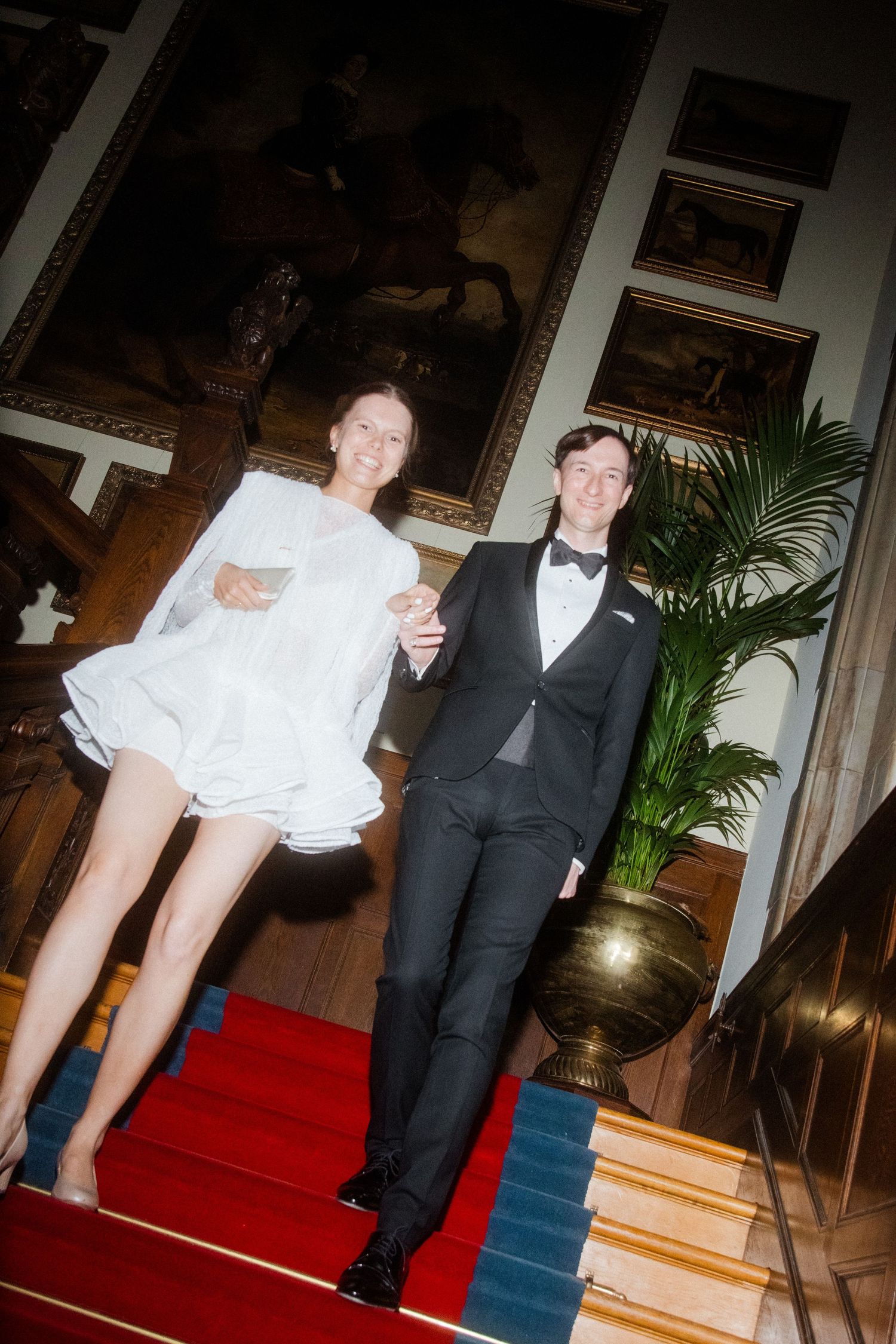 Couple posing playfully on elegant red carpeted stairs in formal attire.