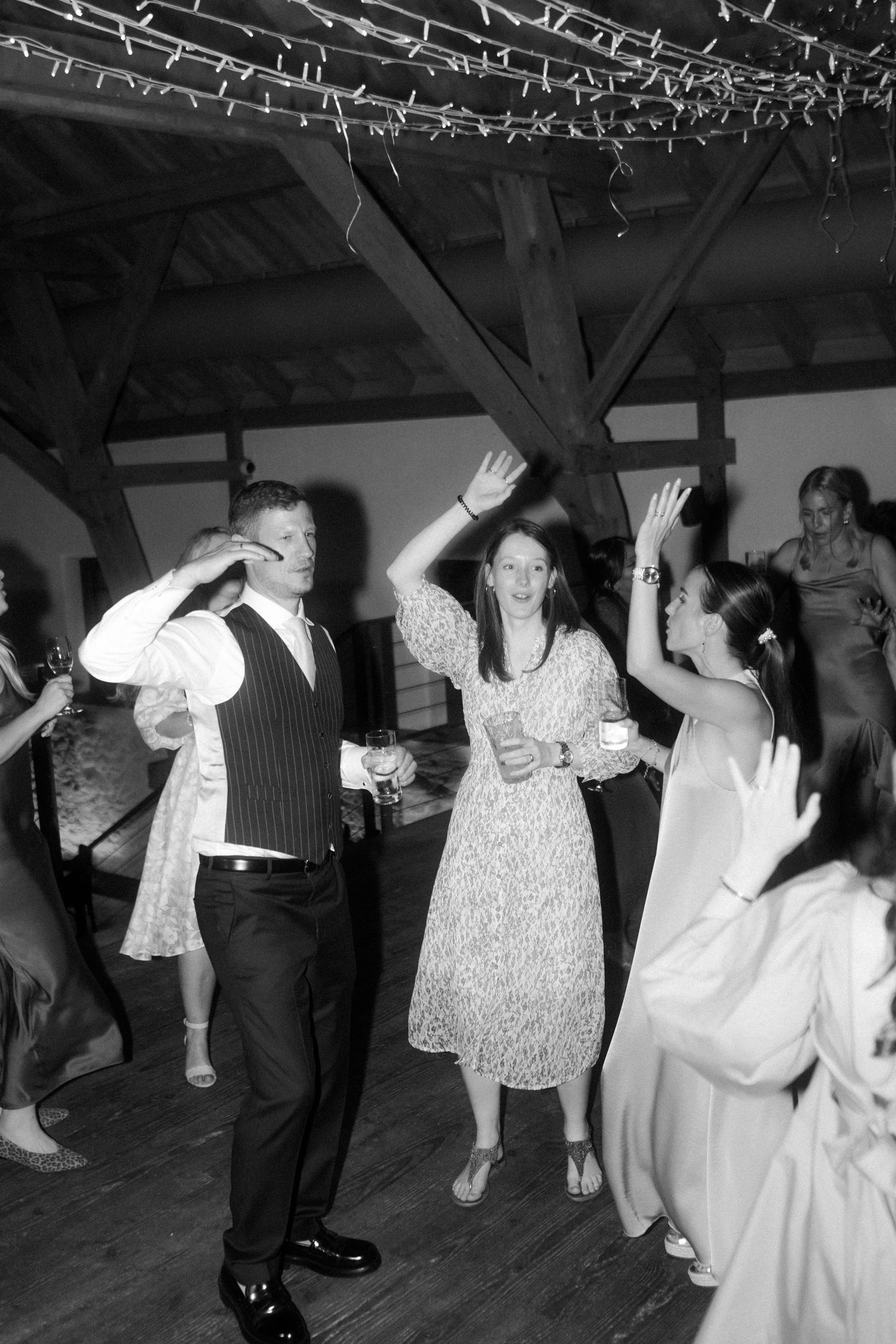 People dancing enthusiastically under string lights at a rustic venue in black and white.