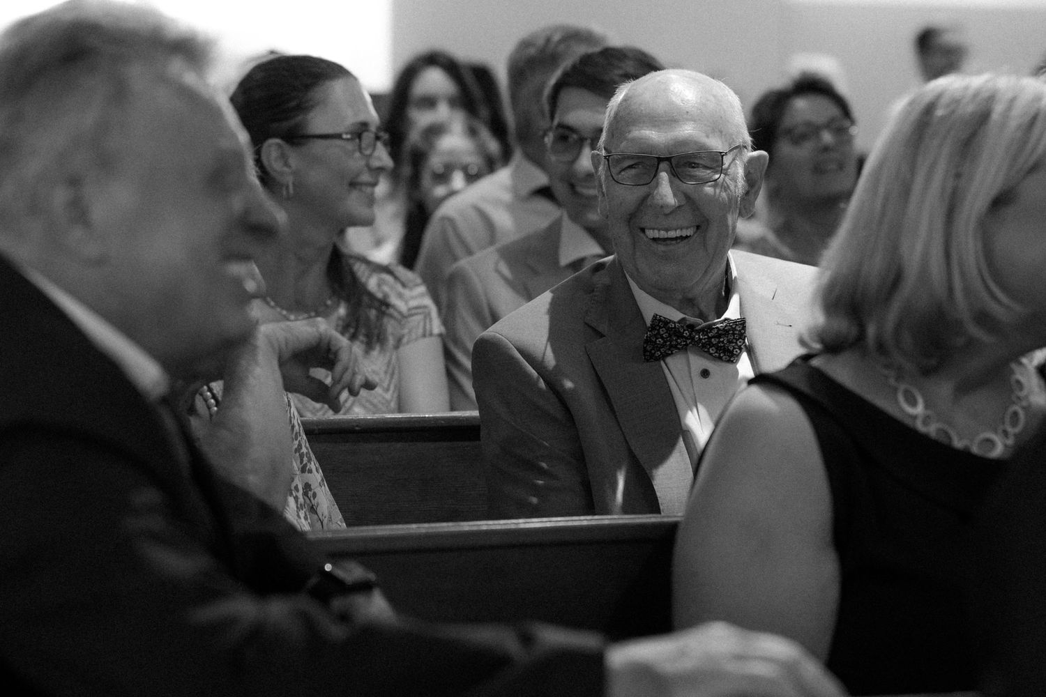 A black and white candid photo of wedding guests smiling and enjoying the ceremony from church pews.