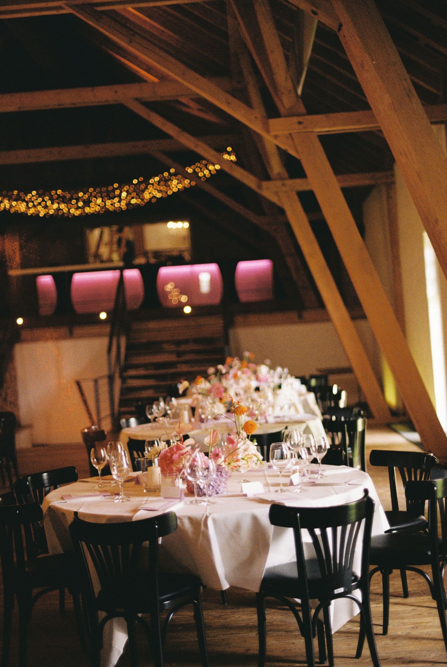 Wedding reception tables set with floral centerpieces under rustic wooden beams and string lights.