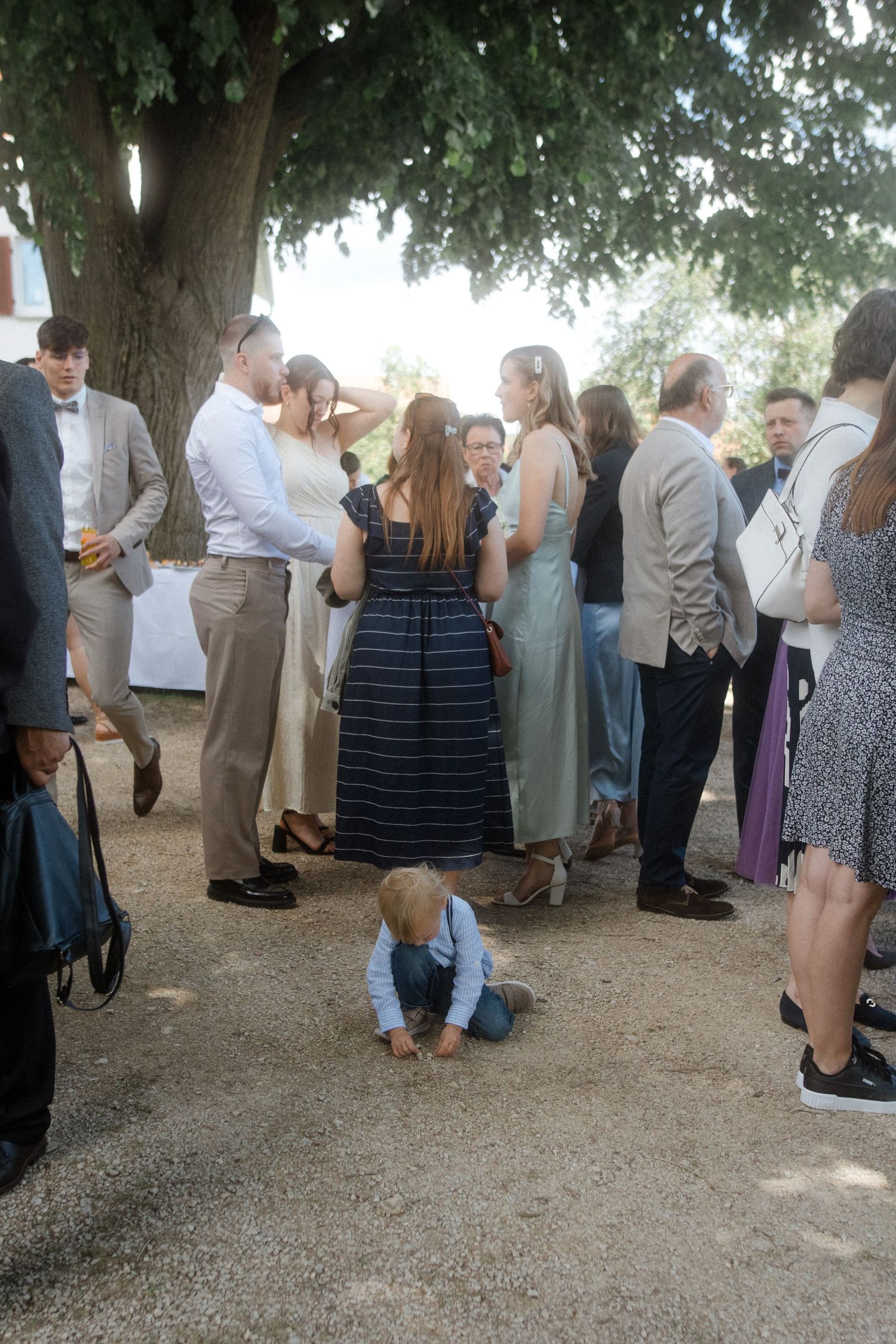 Wedding guests mingling outdoors while small child plays on ground.