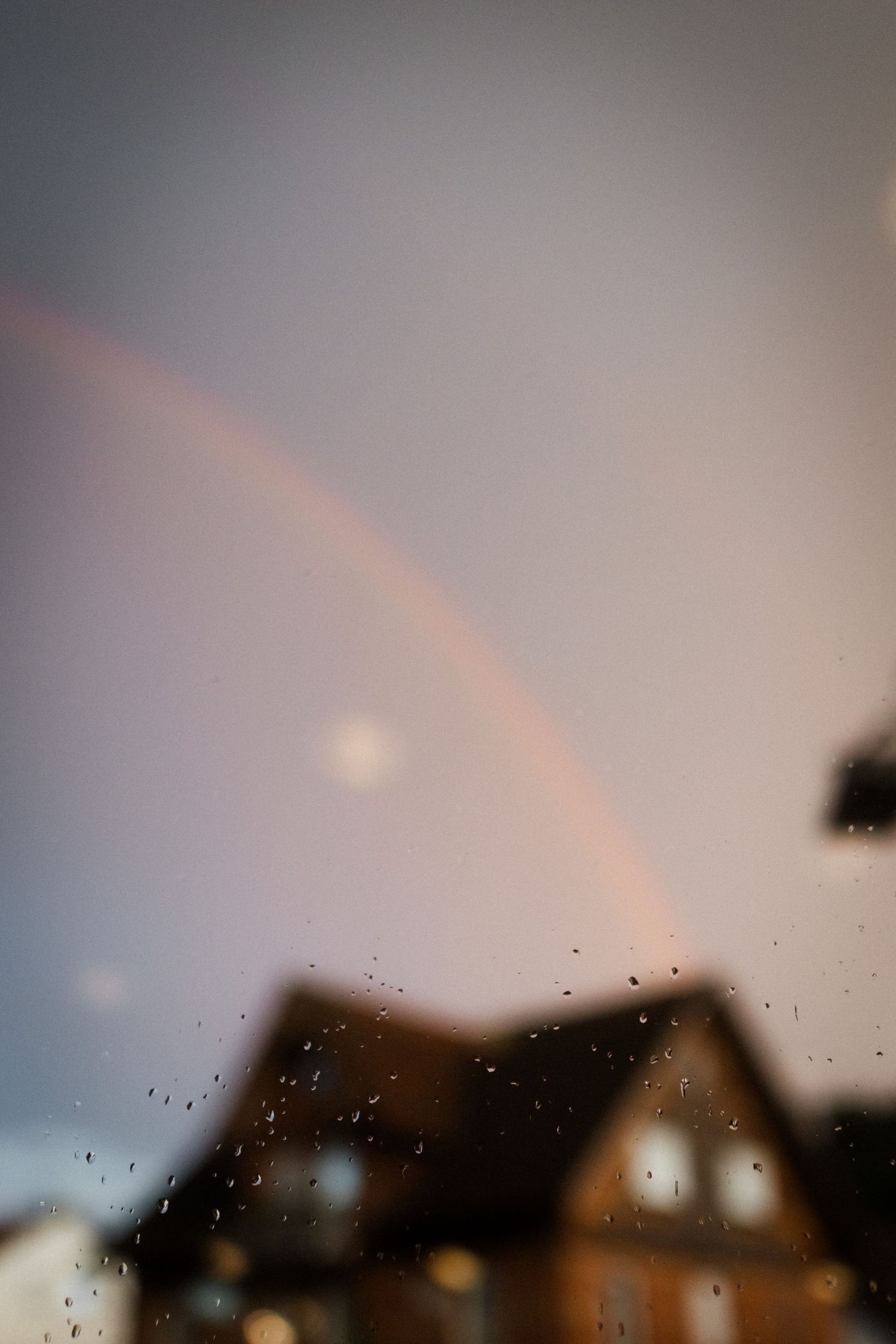 Dreamy rainbow arches over a house silhouette during golden hour with raindrops on window.