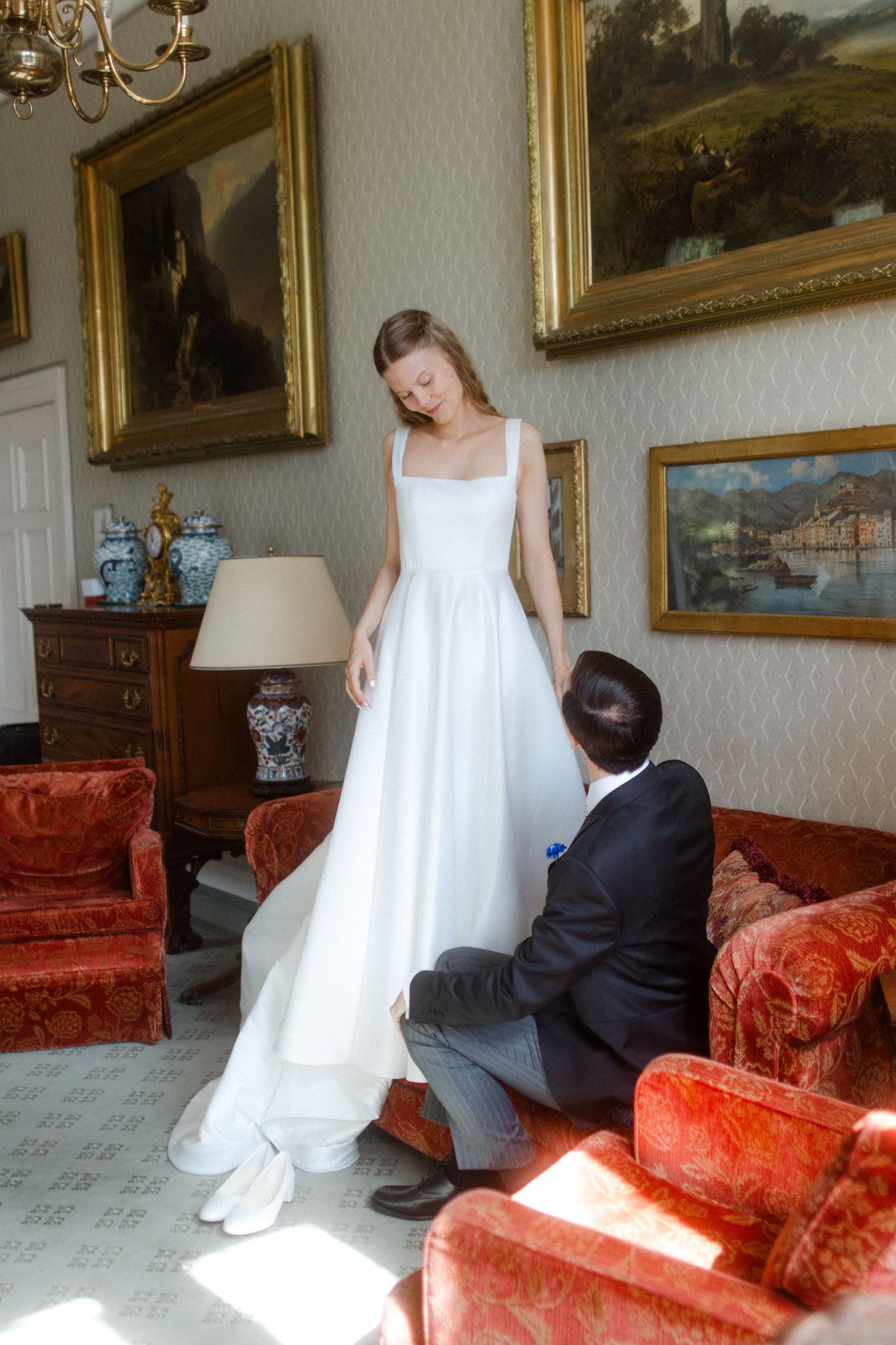 A wedding dress fitting scene in a luxurious room decorated with classical paintings and red velvet furniture.
