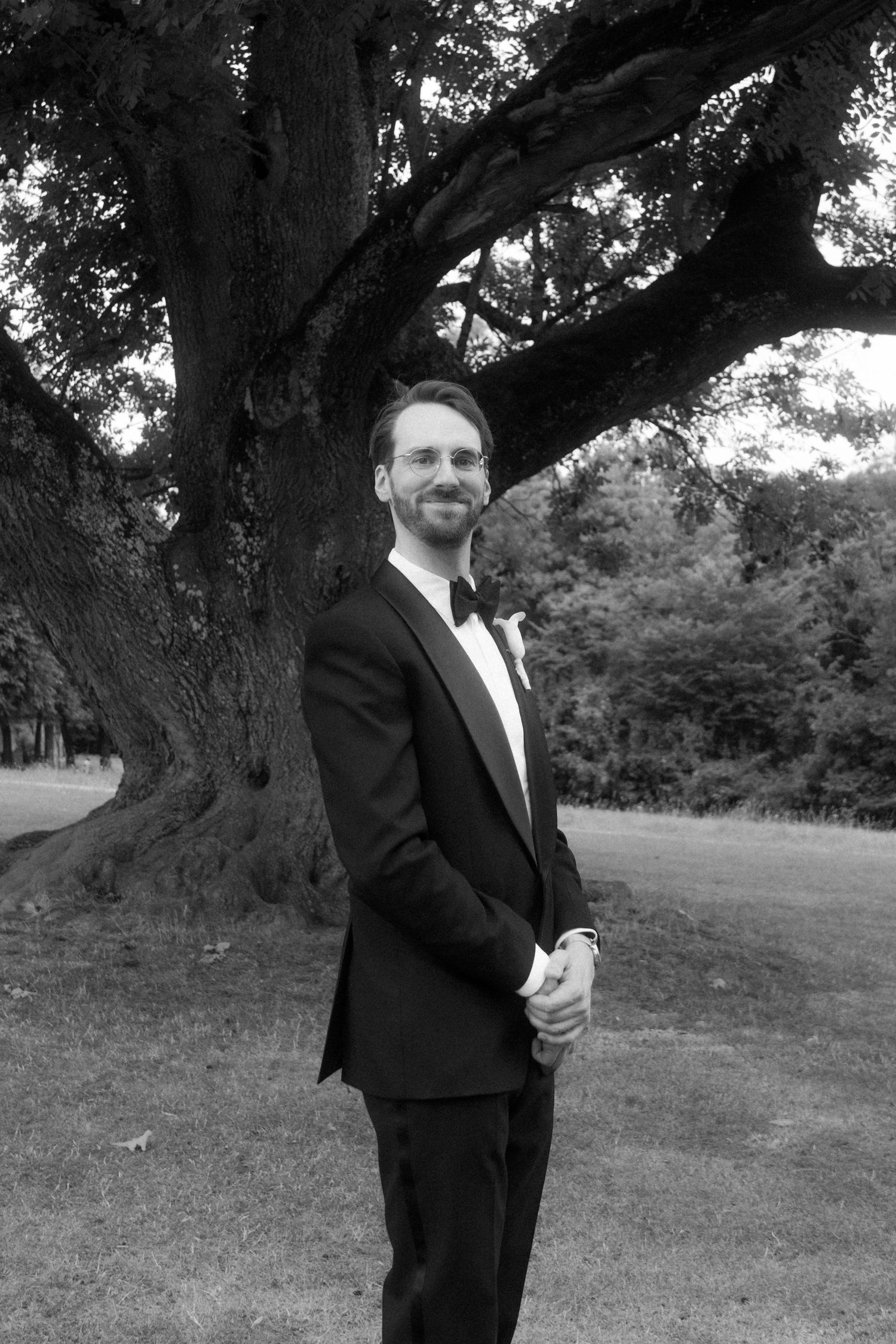 Person in formal attire standing beneath a large tree in black and white photography.