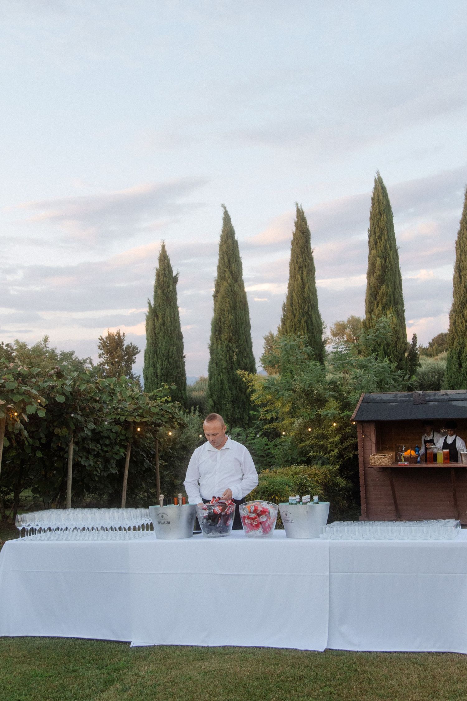 Long white buffet table setup with cypress trees in the background at dusk.