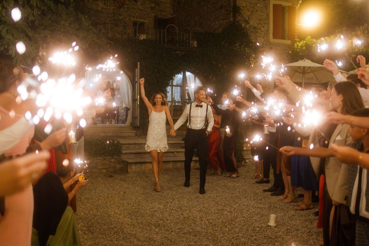 Wedding guests hold sparklers creating a magical light tunnel at night.
