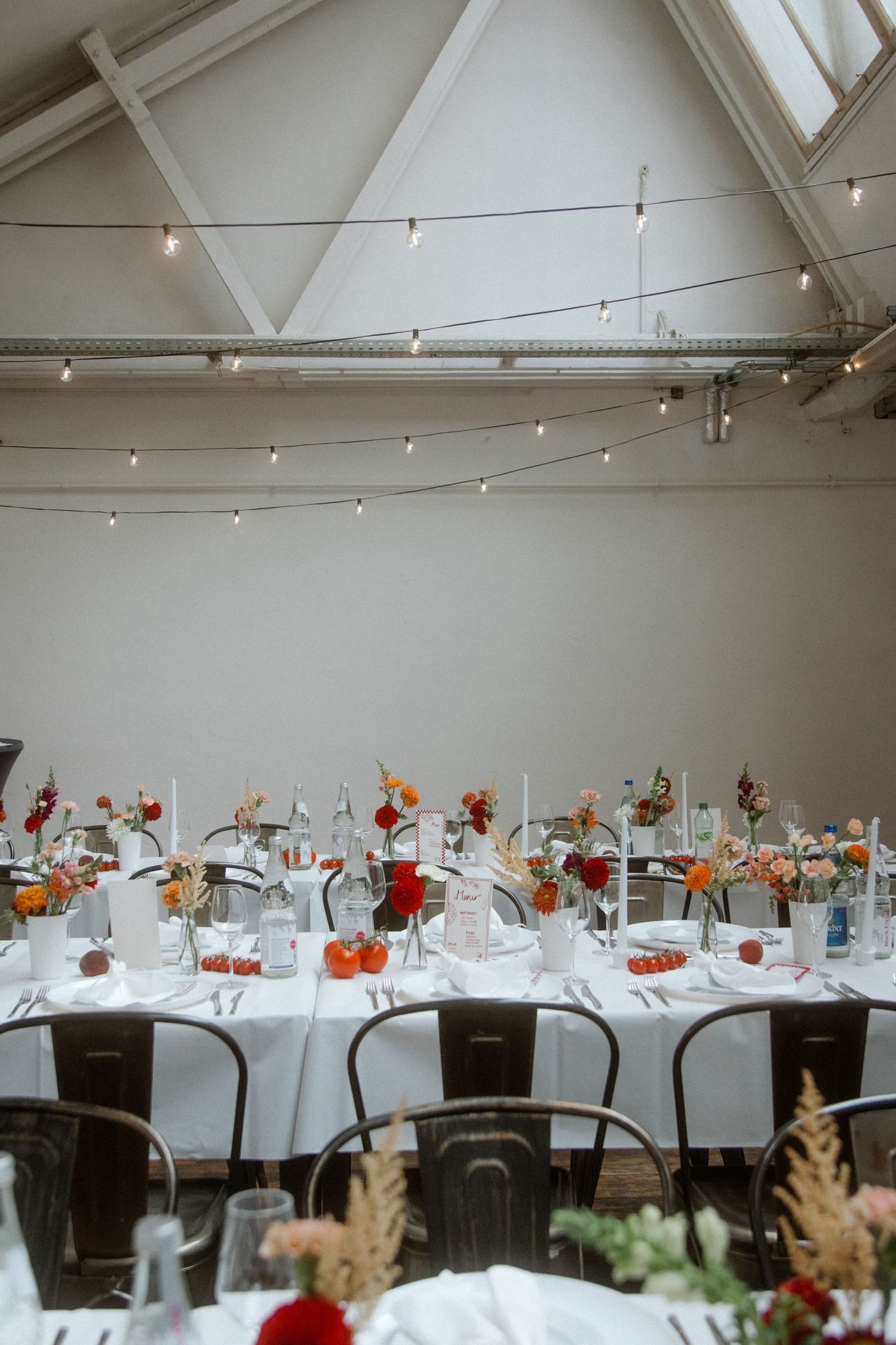 Elegant wedding reception hall with long white tables, colorful floral centerpieces, string lights, and industrial metal chairs.