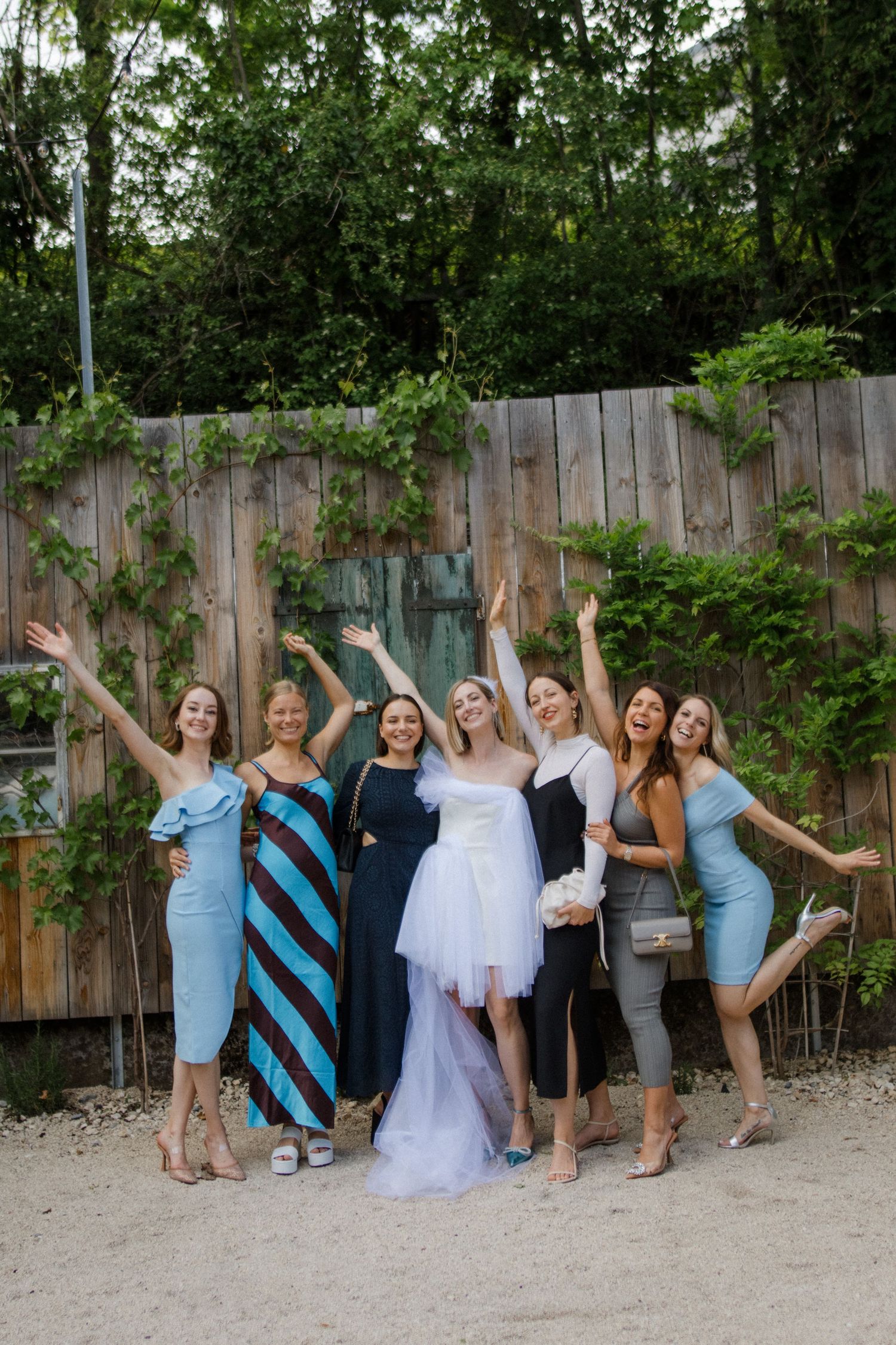 Wedding party striking fun poses together on a sandy ground against a wooden fence.