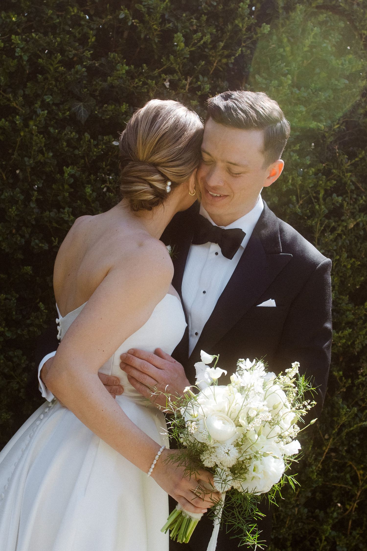Bride and groom embrace outdoors, she holds white bouquet, he wears black tuxedo with bow tie, greenery background.
