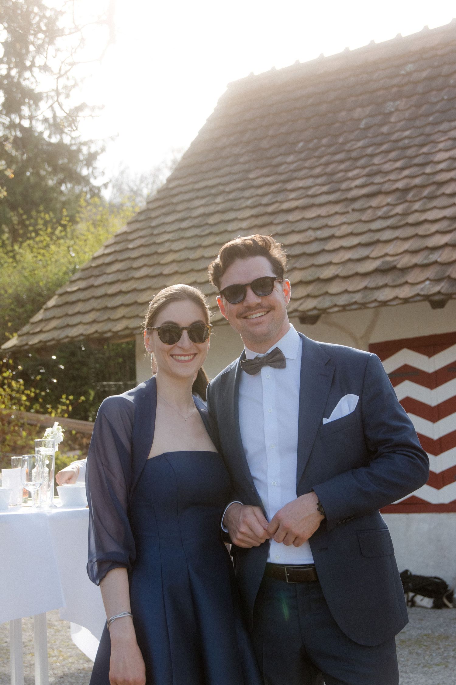 Couple in matching navy blue outfits and sunglasses posing outdoors at a wedding reception near a rustic building.