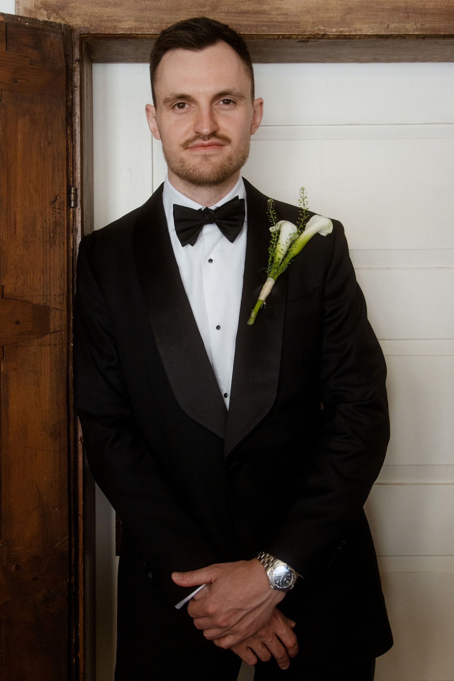 Groom in black tuxedo with bow tie and white boutonniere poses confidently before wooden doors.