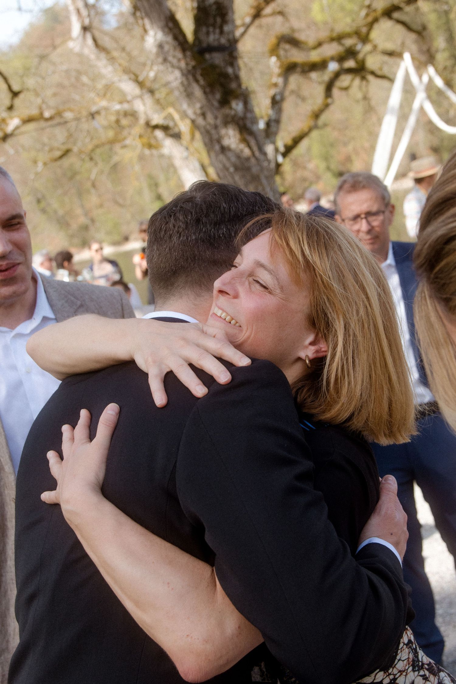 Woman smiling joyfully while hugging a man in a suit at an outdoor gathering.