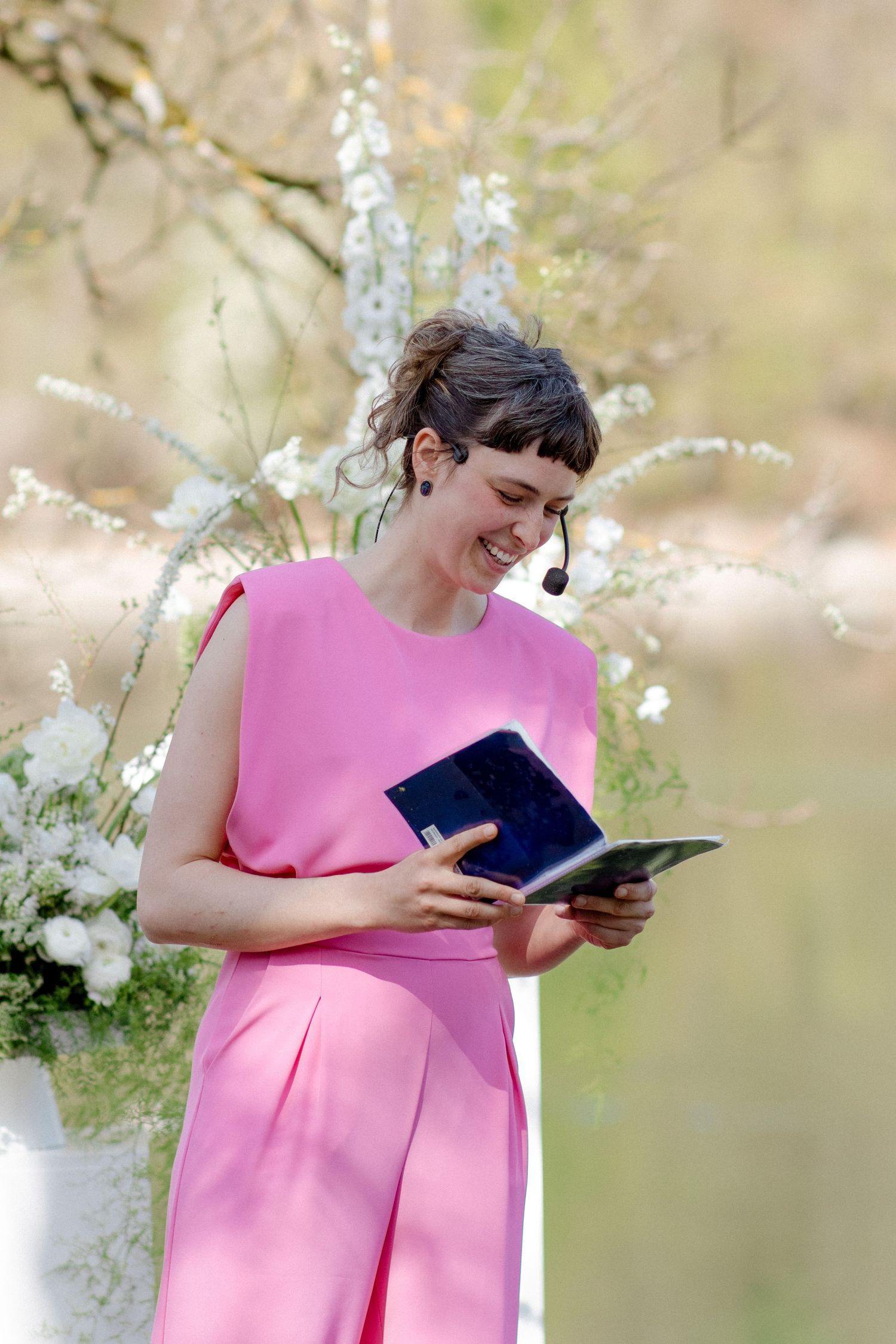 Smiling wedding officiant in a pink dress reads from a book at an outdoor ceremony with white floral decor.