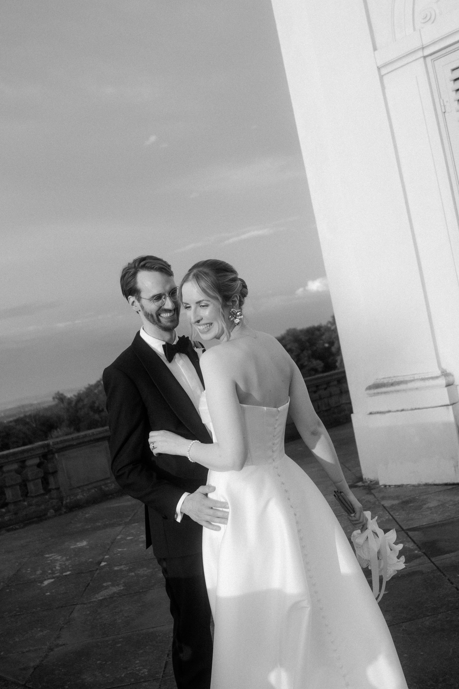 Wedding couple sharing a joyful moment against a white building exterior.