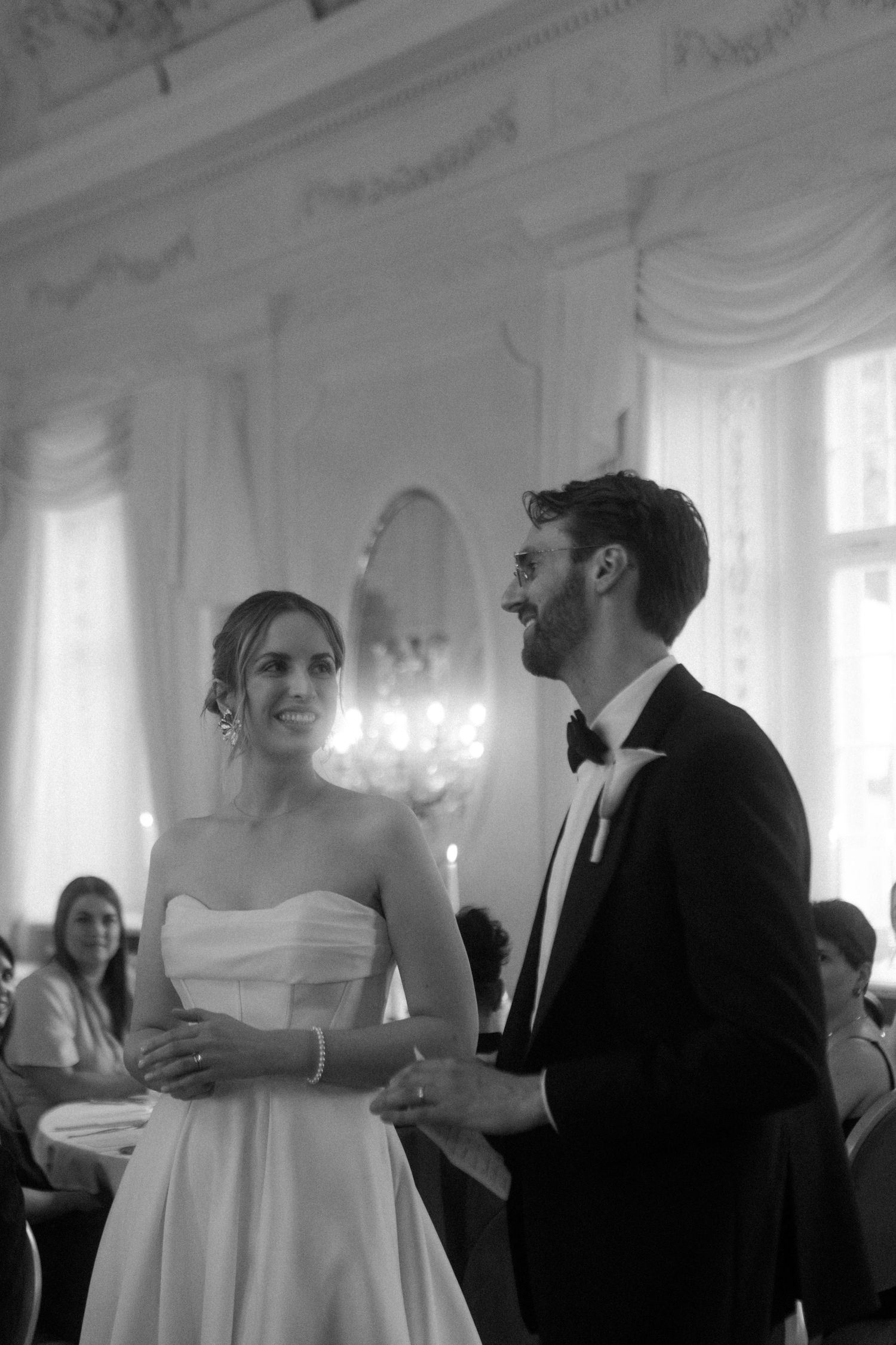 Black and white wedding photo showing guests mingling in an ornate room with chandeliers.