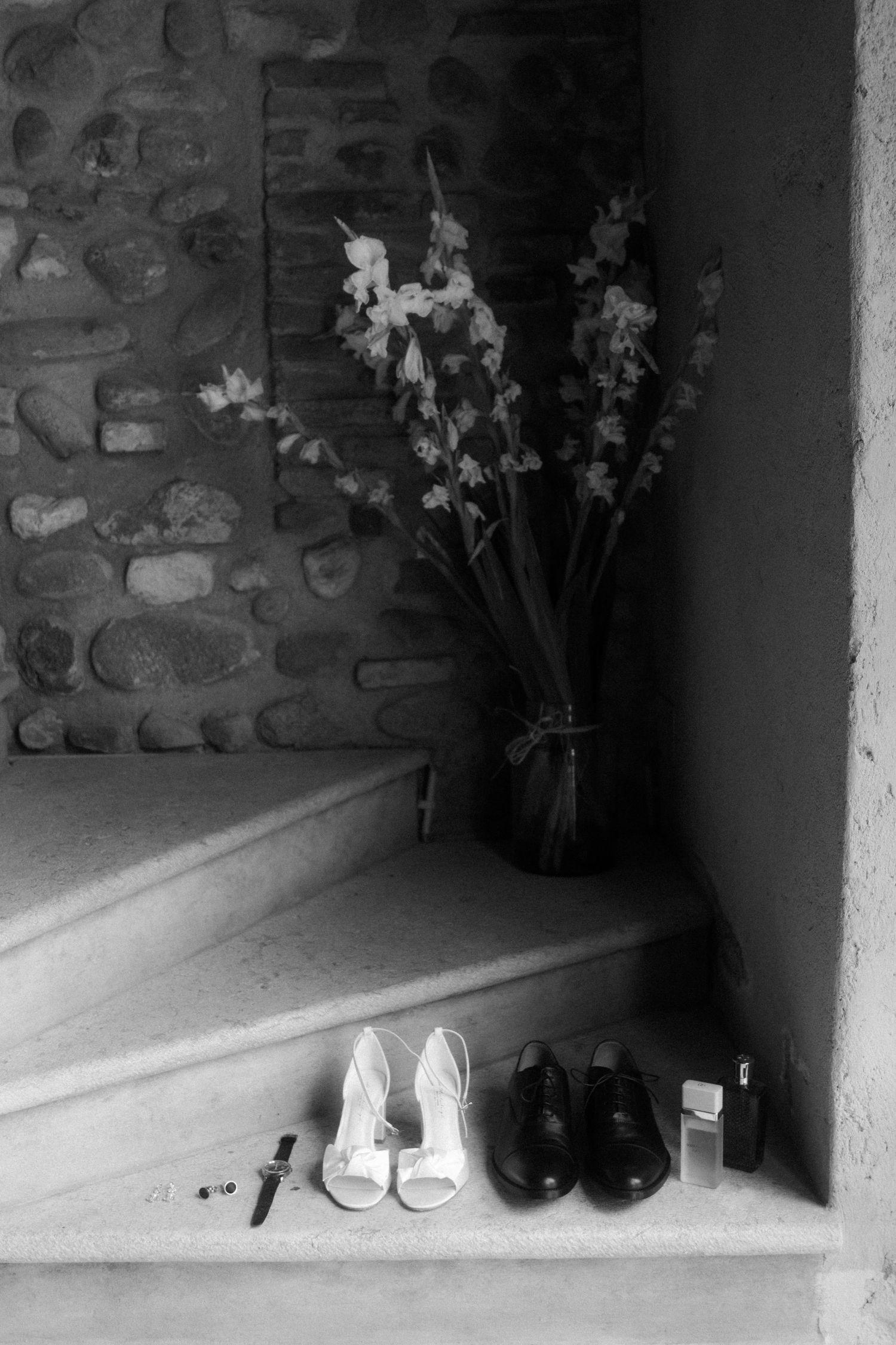 Black and white photo of shoes arranged in alcove with stone walls.