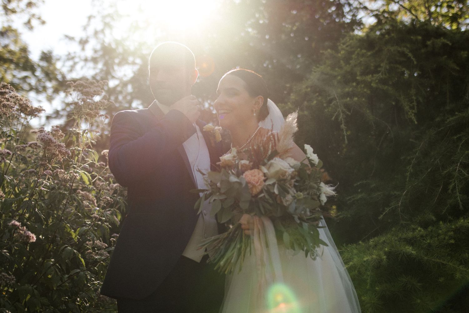 Joyful wedding couple sharing a laugh in a sun-drenched garden setting.