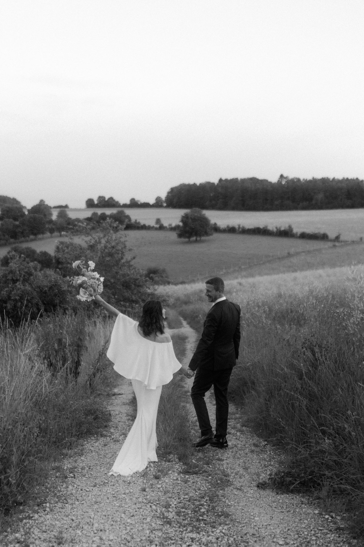 A black and white photo of newlyweds standing on a rural path overlooking rolling hills.