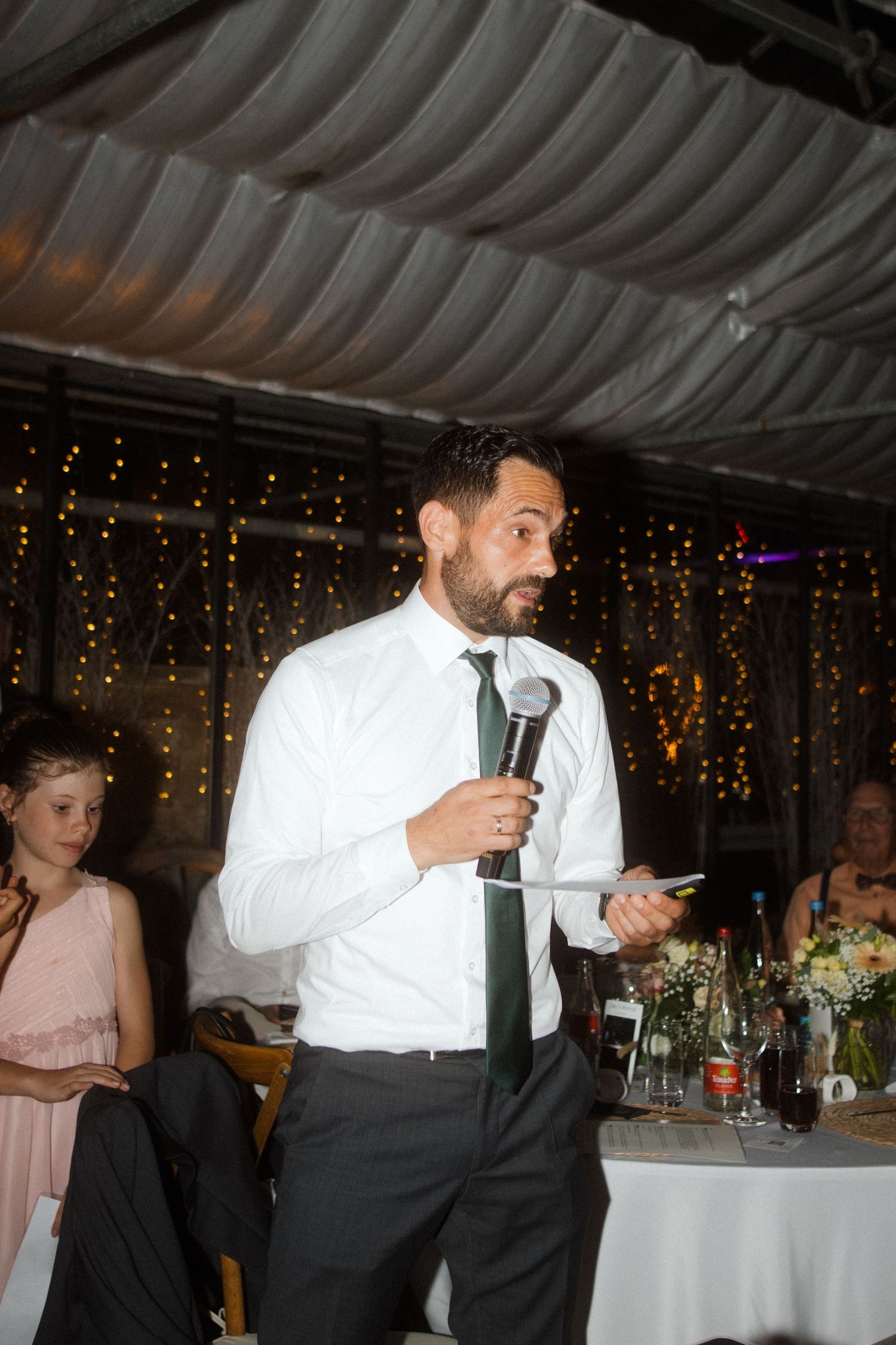 A person in formal attire giving a speech with a microphone at a wedding reception under draped fabric ceiling.