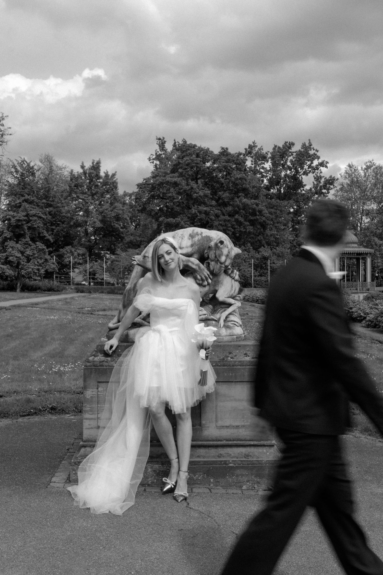 Artistic black and white capture of bride posing by statue as groom walks past.