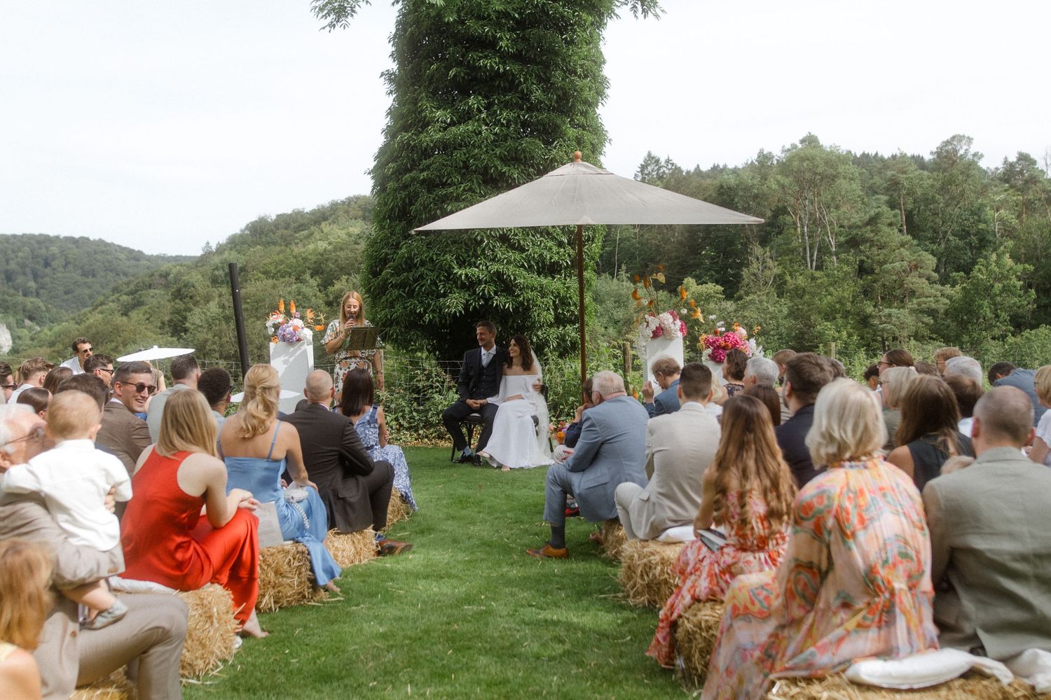 Scenic outdoor wedding ceremony with guests seated under umbrella in natural setting.