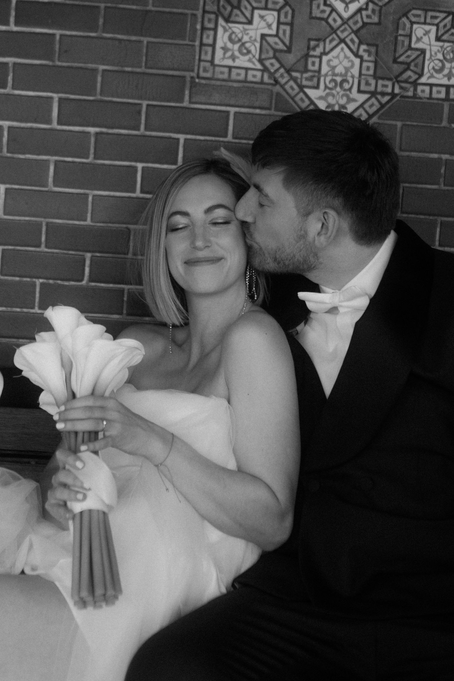 Black and white portrait of wedding couple sharing tender moment on bench with calla lily bouquet.