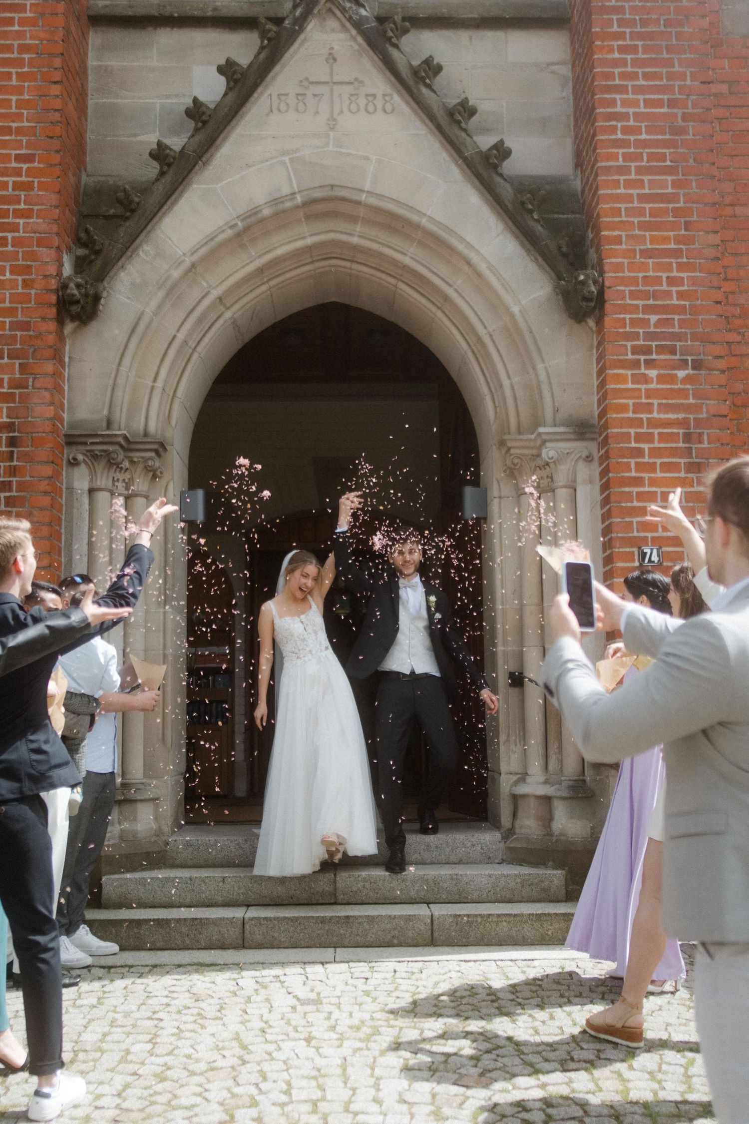 Newlyweds exit church while guests throw confetti in afternoon sunlight.