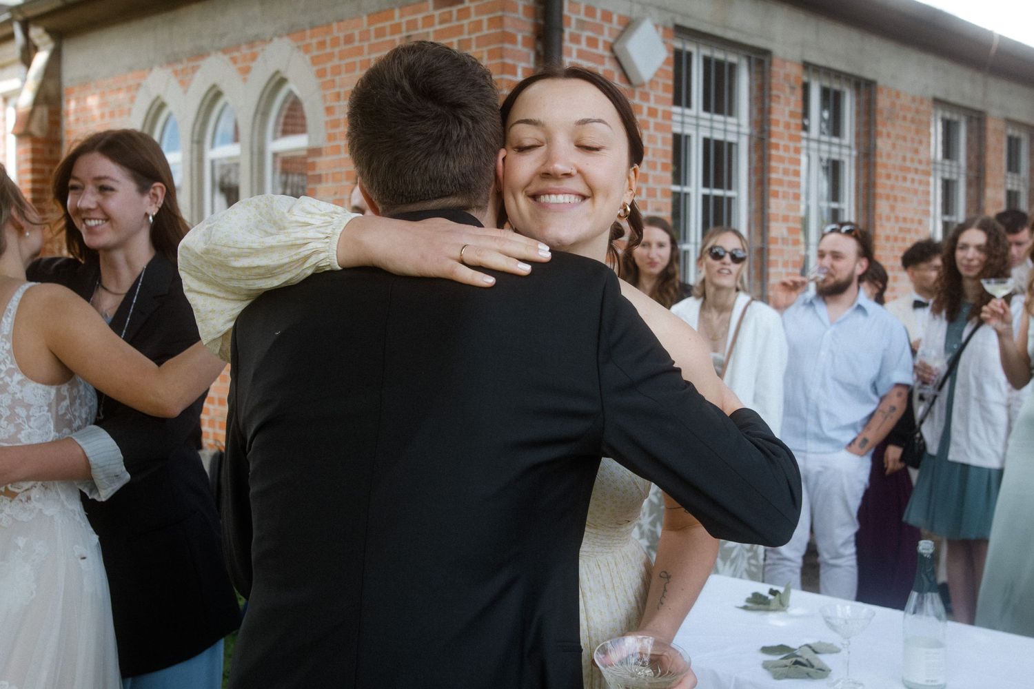 Joyful embrace at an outdoor wedding reception with guests looking on near a brick building.