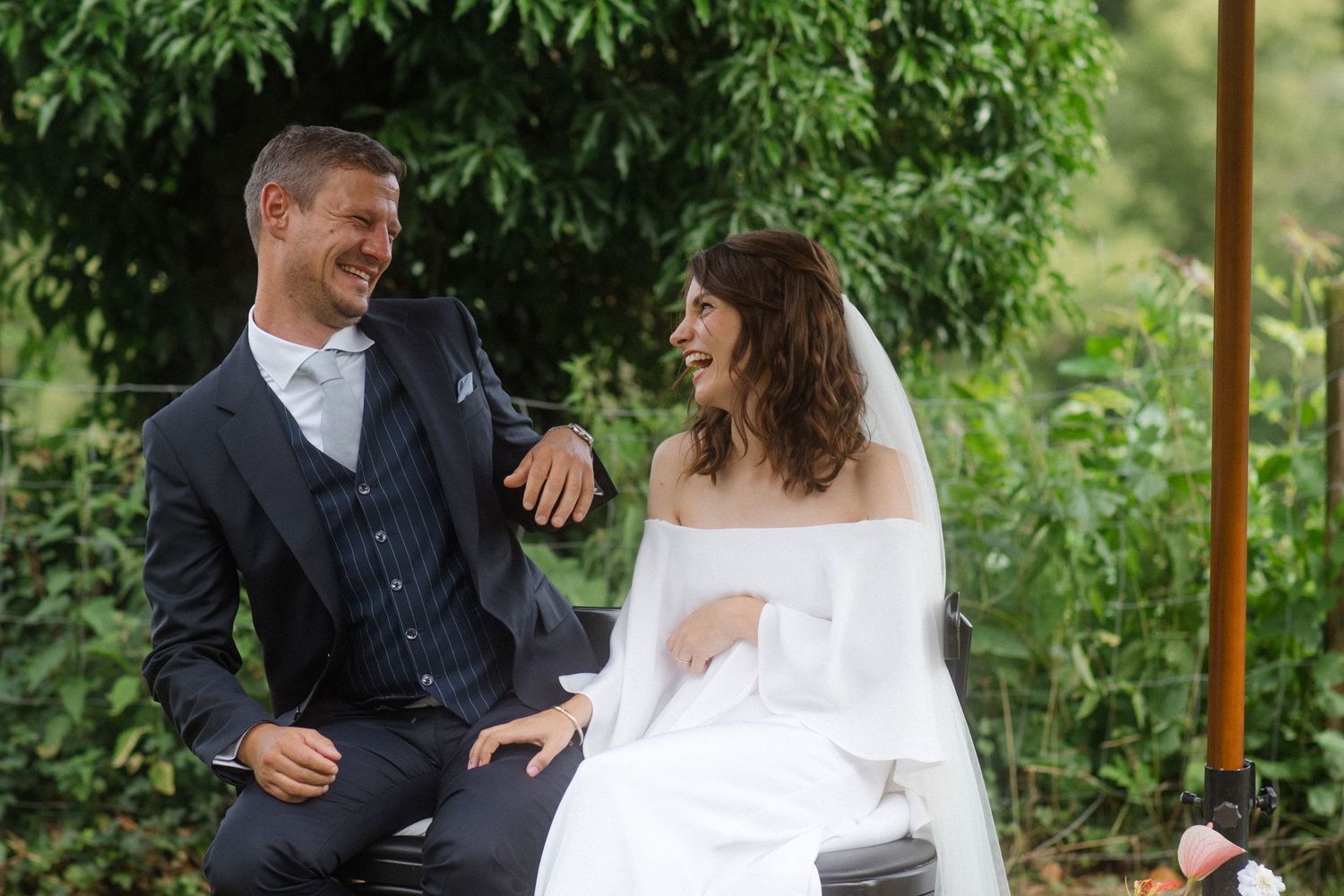 A couple shares a laugh while sitting outdoors during their garden wedding ceremony.