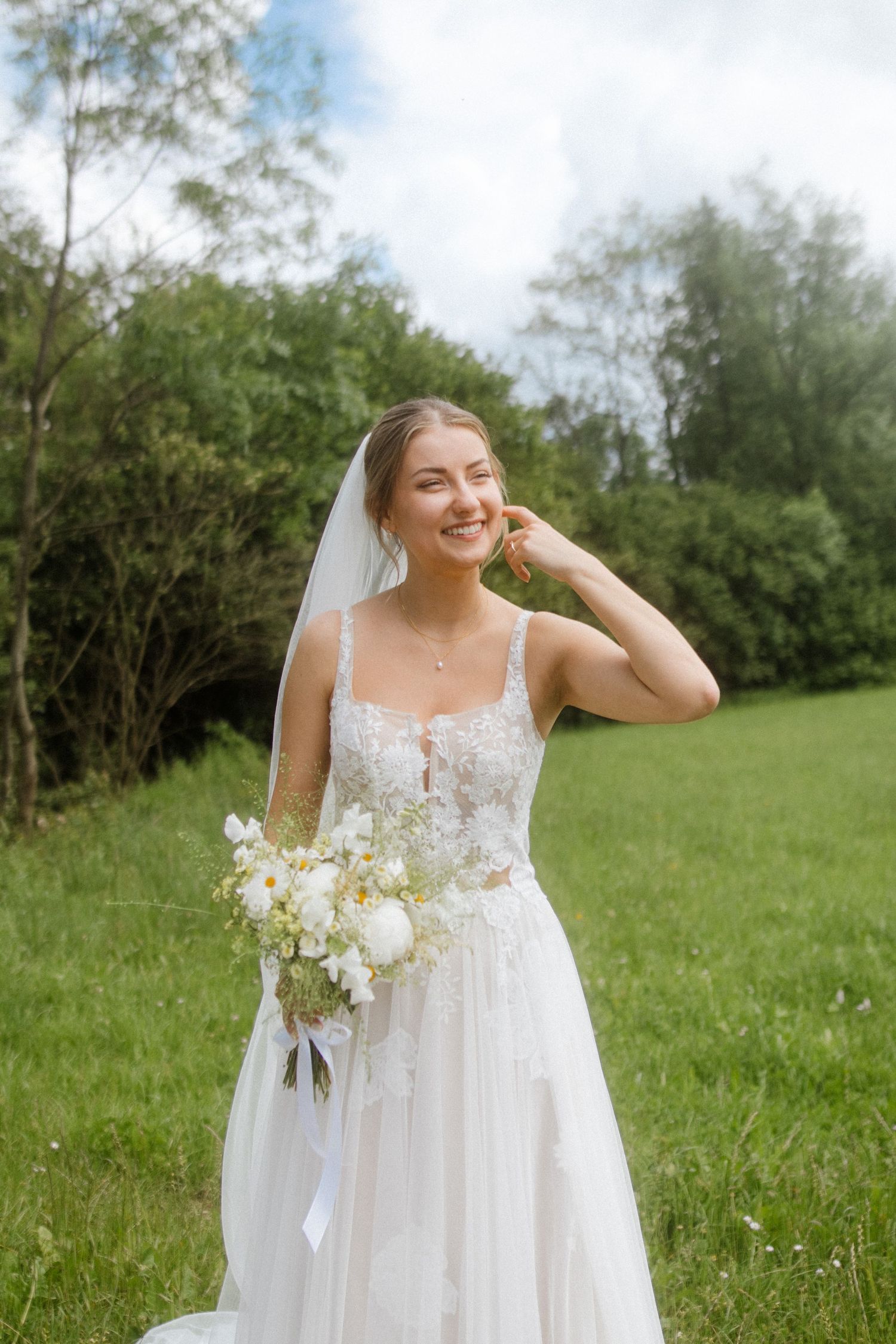 Bride in white lace dress laughs joyfully while holding bouquet in outdoor garden setting.