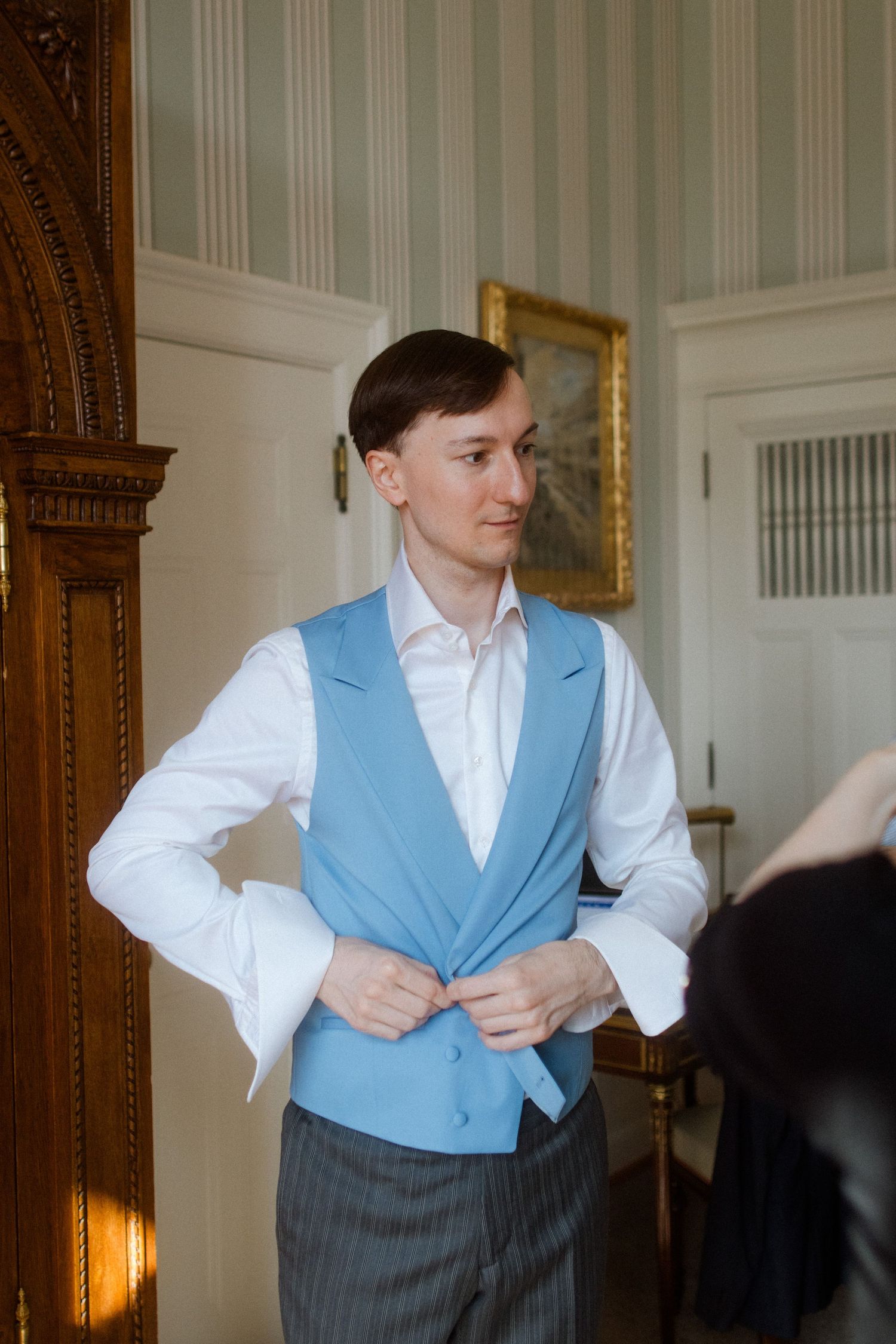 Person wearing light blue vest and white shirt getting dressed in elegant room.