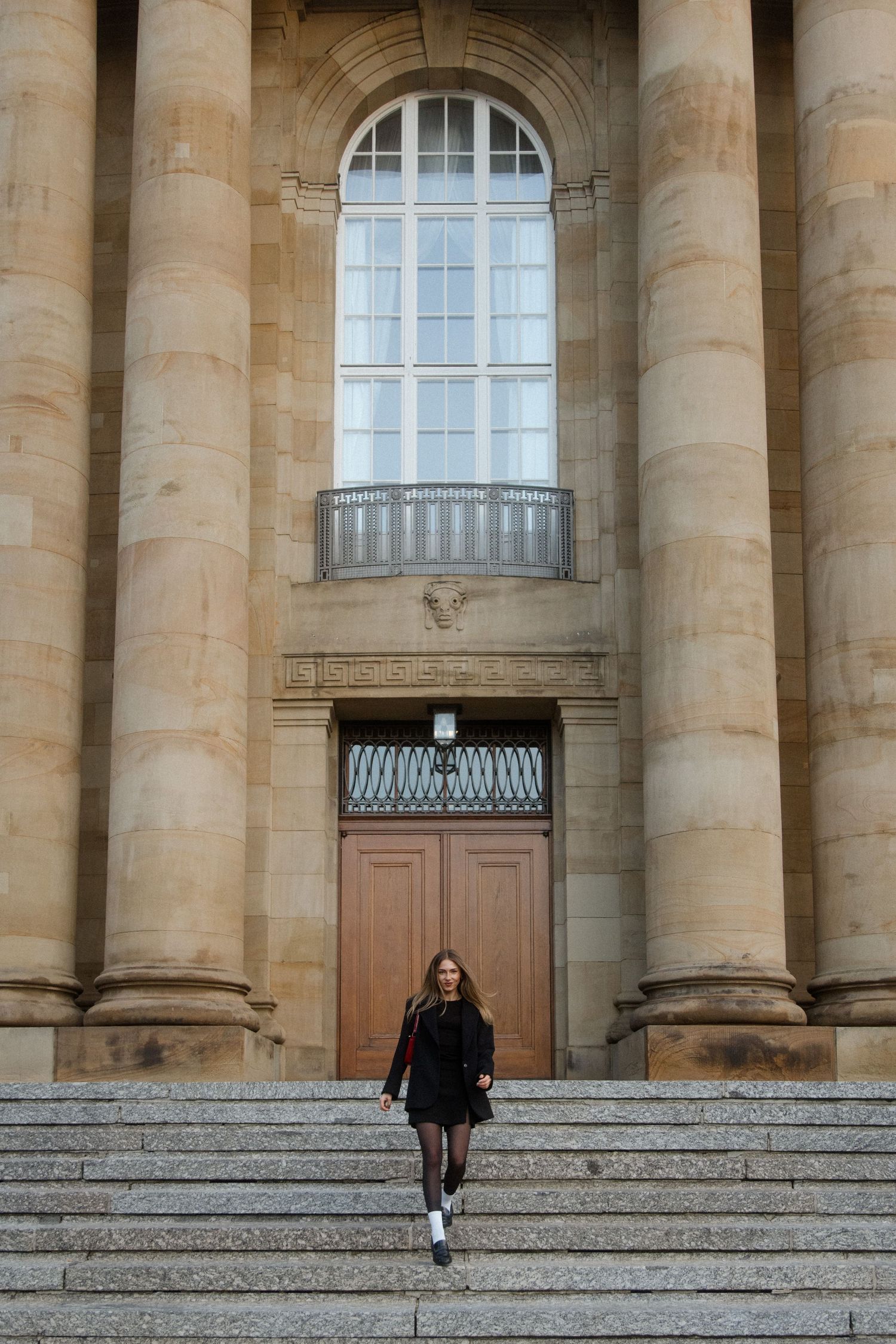 Person sits on stone steps in front of a grand neoclassical building with tall columns and wooden doors.