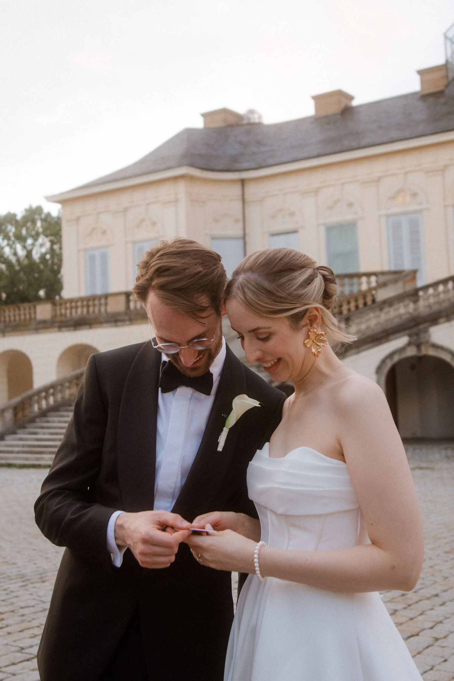 Couple in wedding attire examining rings outside an elegant stone building.