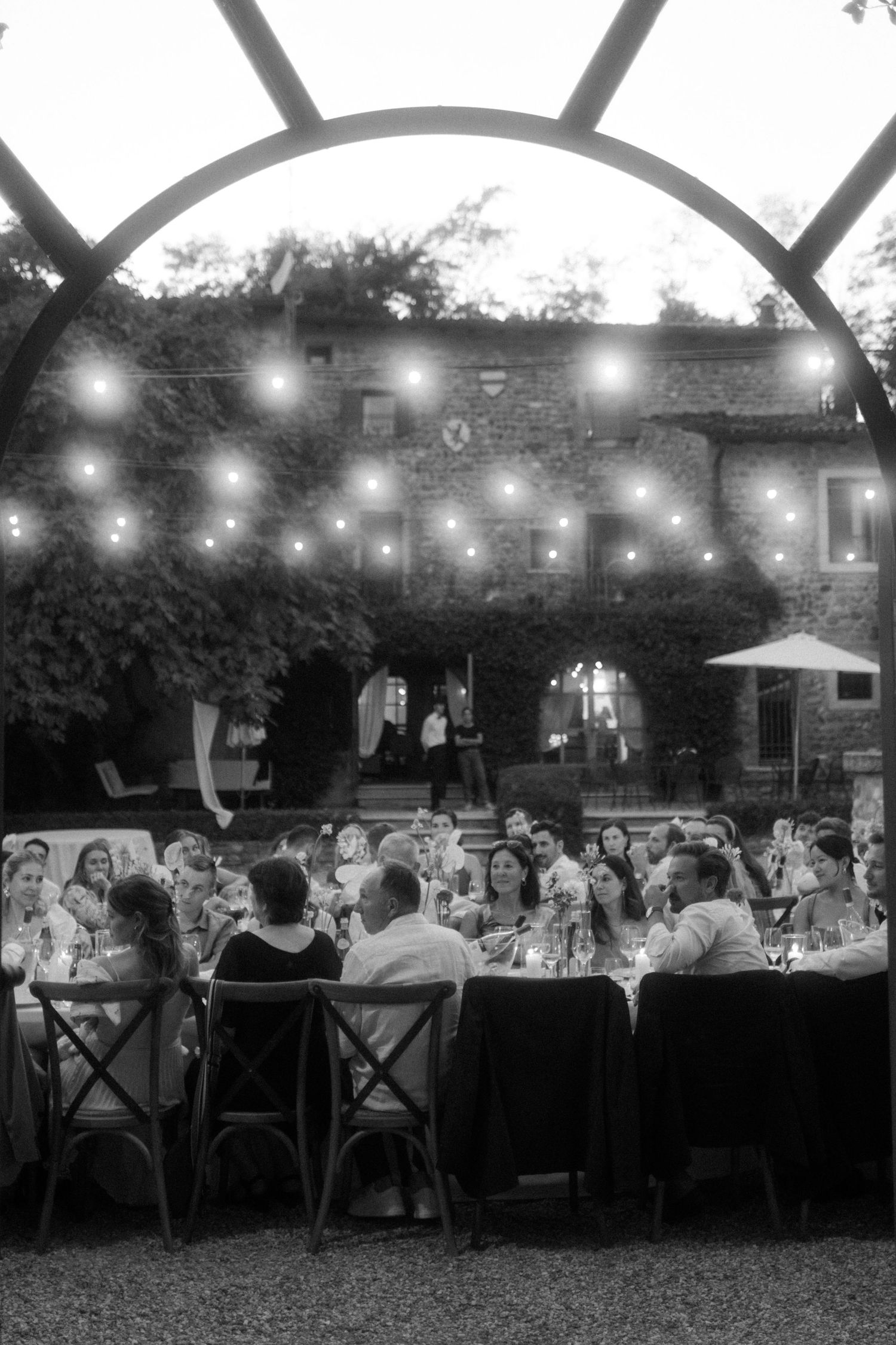 Outdoor evening reception under string lights with guests seated at long dining tables.