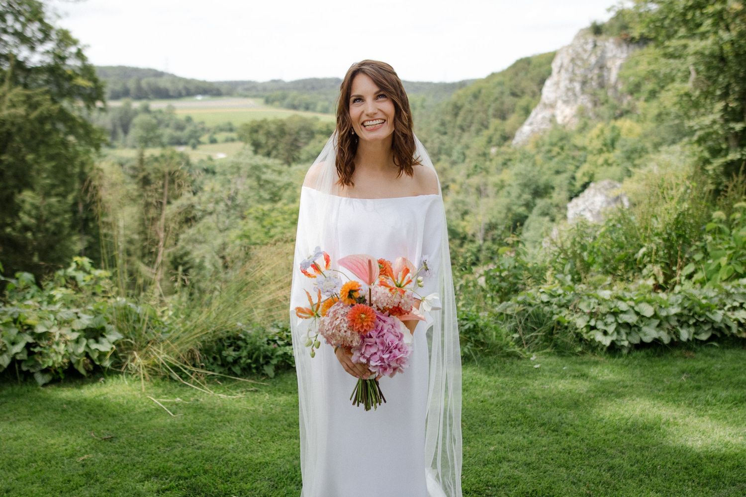 A bride stands on a grassy overlook holding a colorful bouquet with rolling hills in the background.