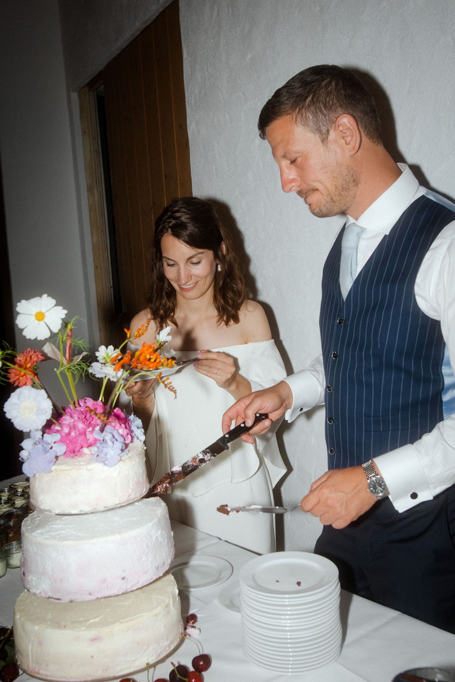A newlywed couple serves their white tiered wedding cake decorated with vibrant flowers.