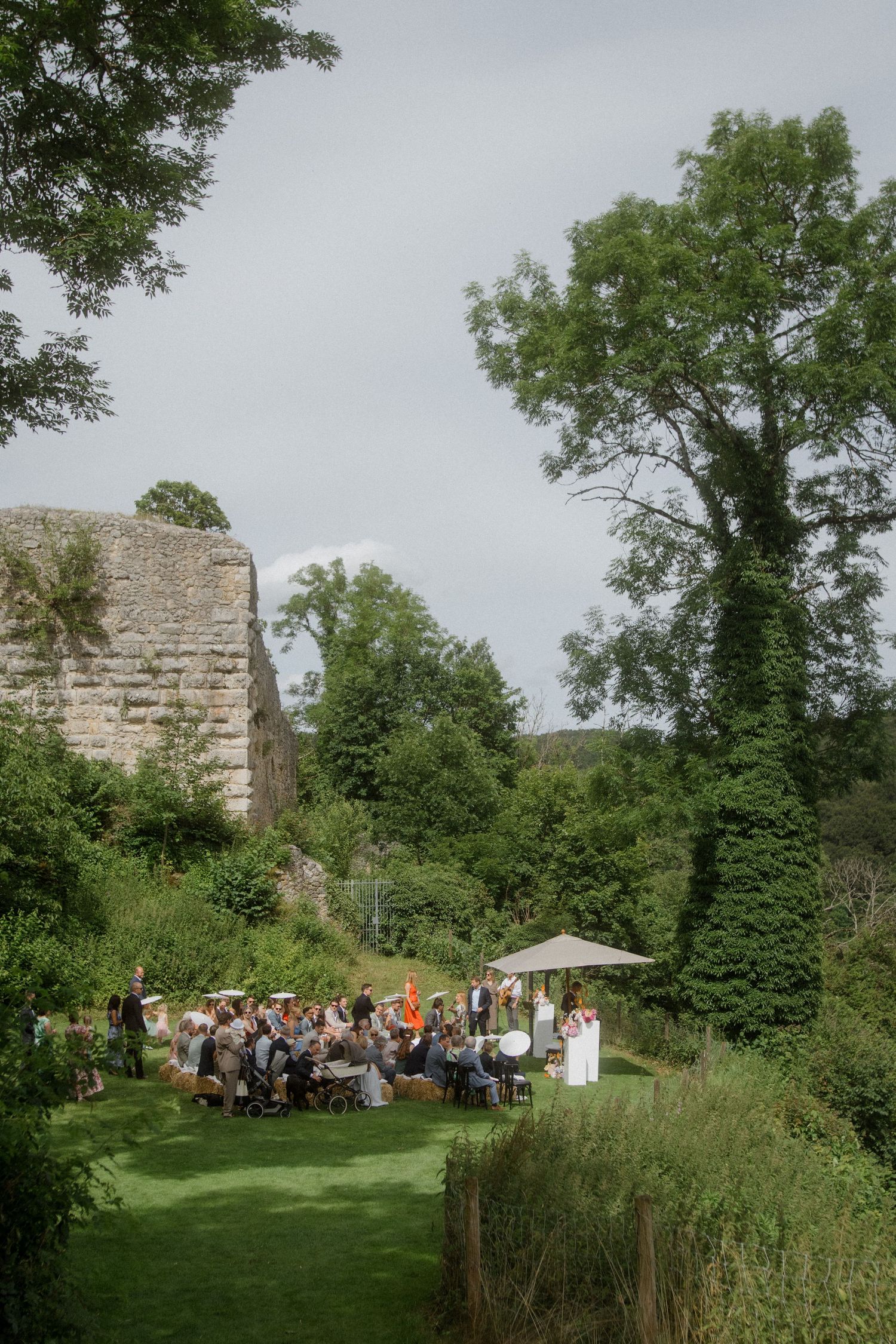Garden wedding ceremony with guests gathered under stone walls and trees.