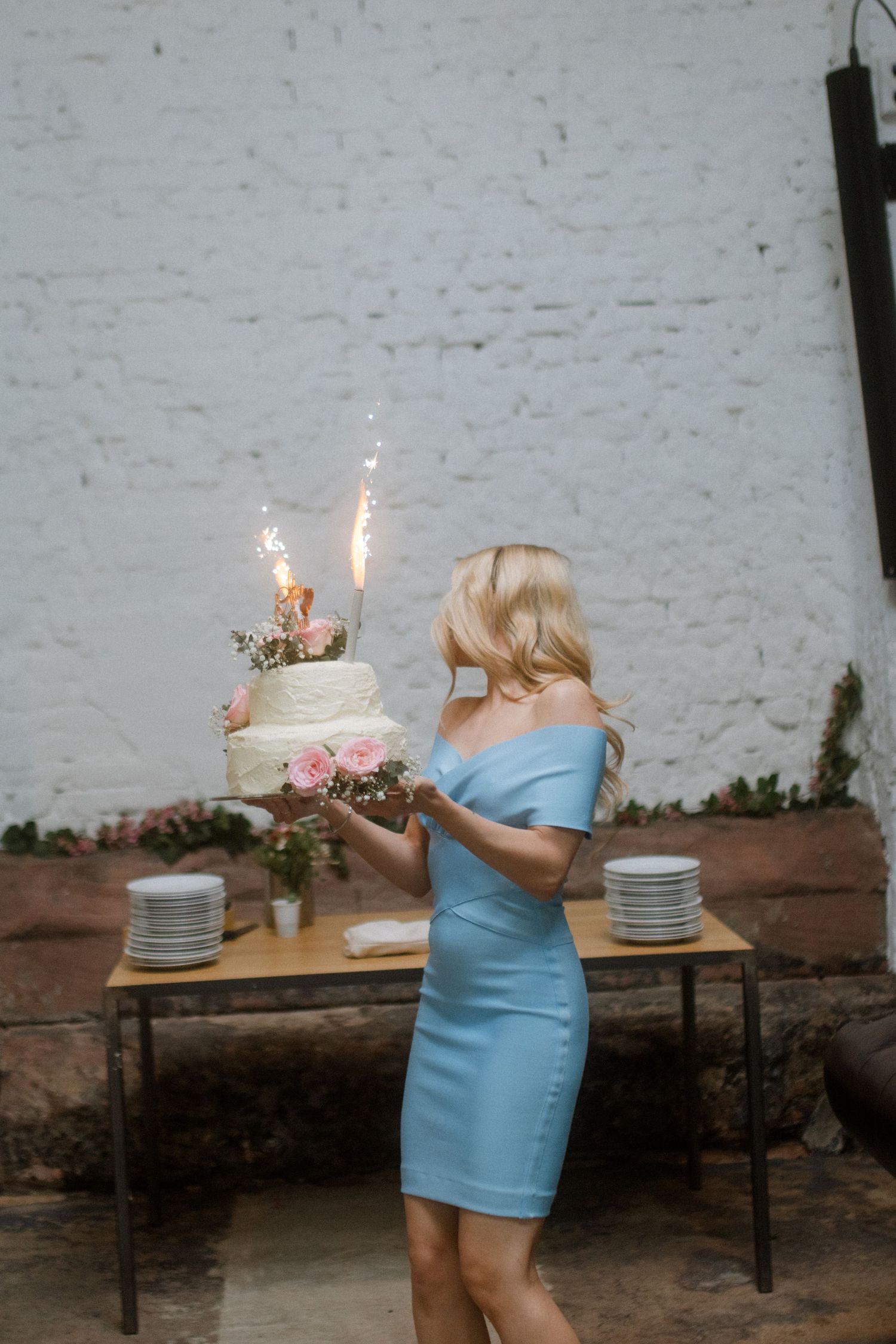 A person in a blue dress holds a wedding cake with sparklers against a white brick wall.