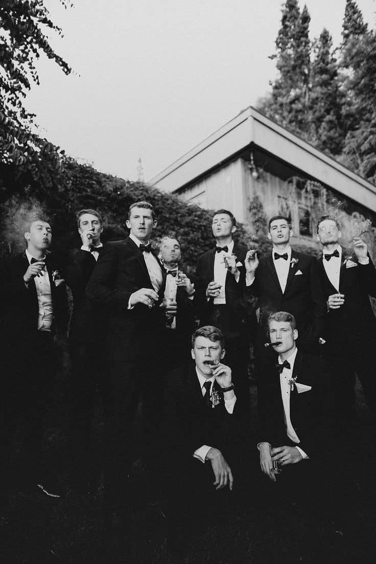 Vintage black and white photo of groomsmen in tuxedos drinking beverages outside a building.