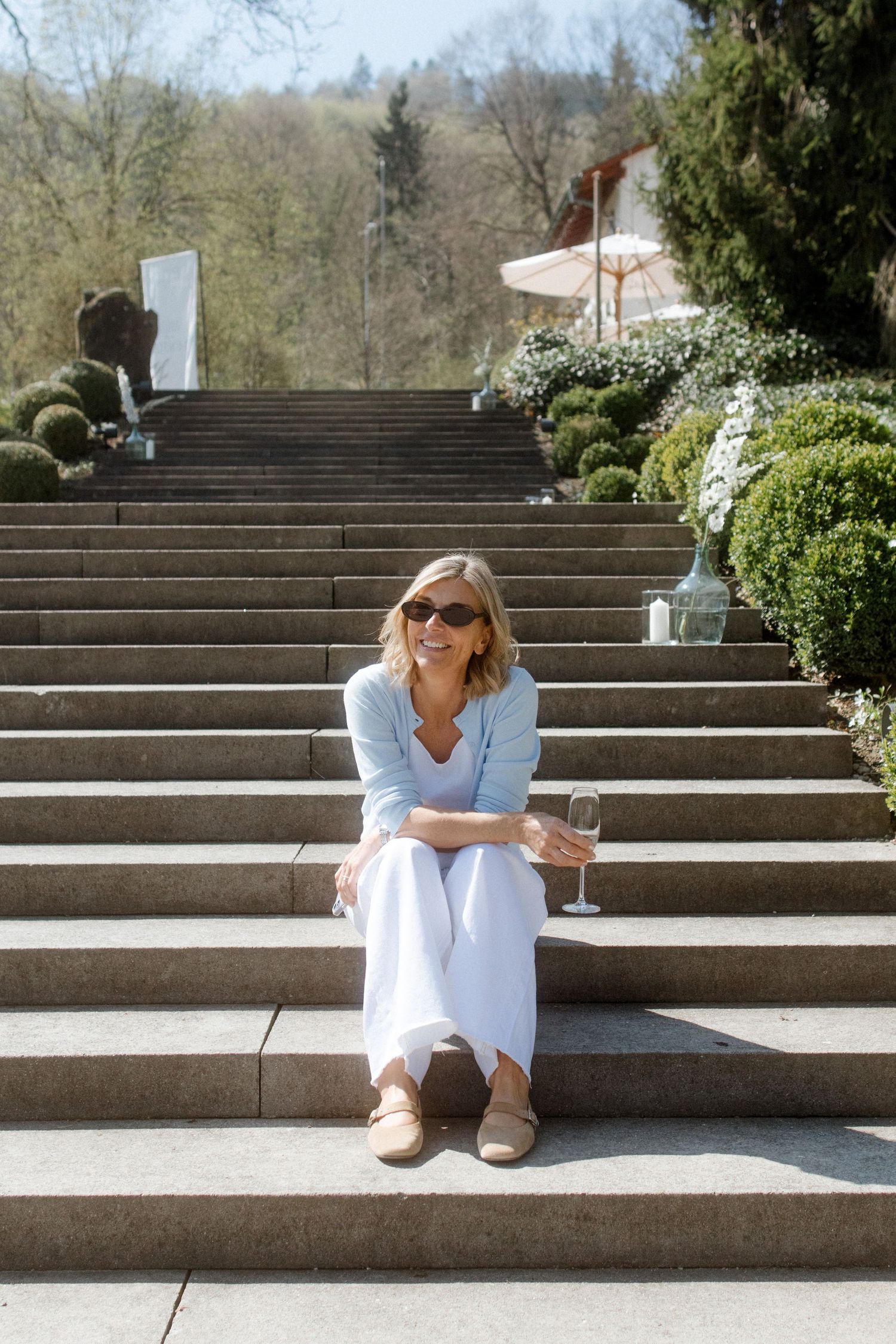 Woman in light blue blazer and white pants sitting on stone steps holding a glass of white wine outdoors.