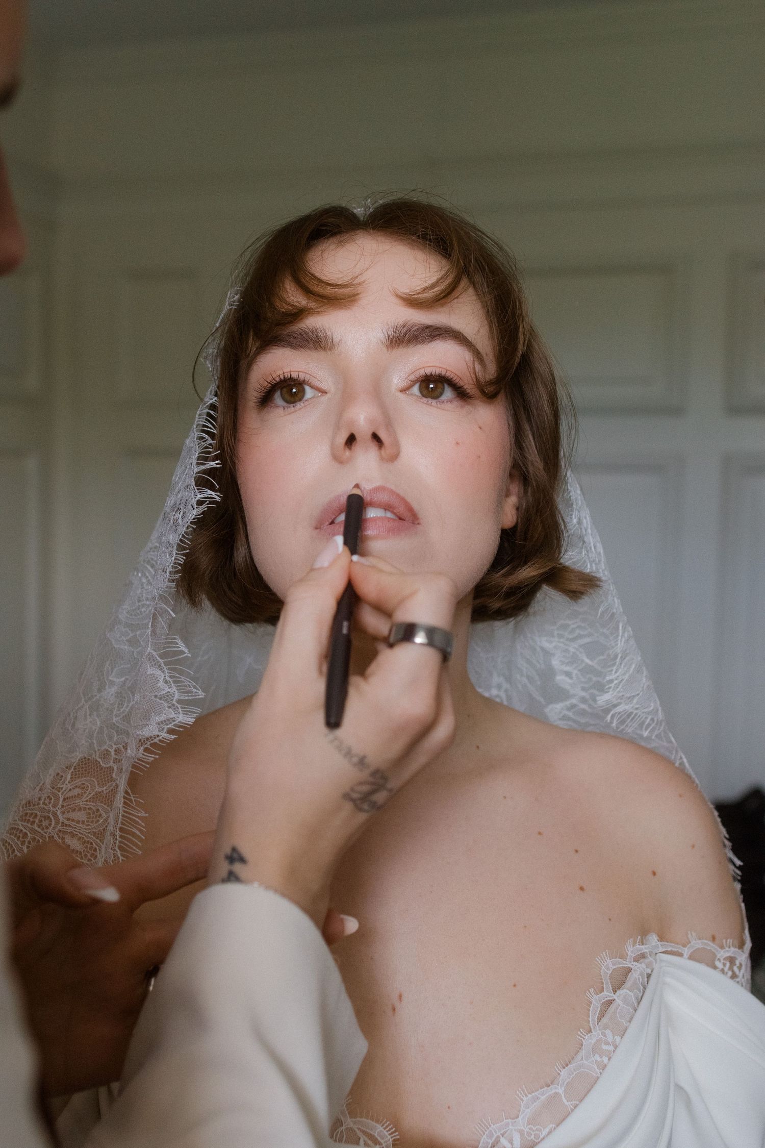 Bride in white lace veil getting lipstick applied by makeup artist during bridal preparation.