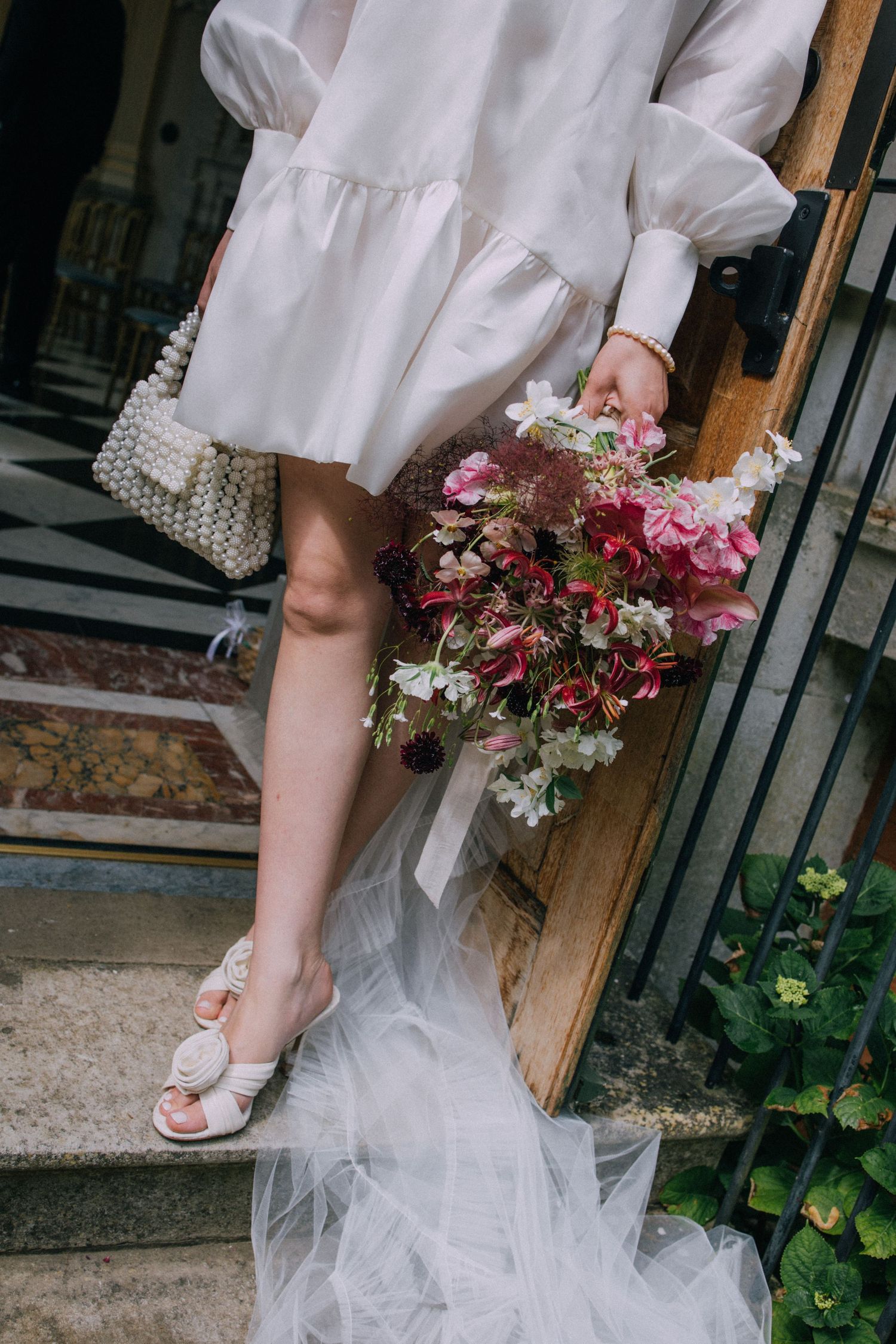 Bride in white mini dress holding pink floral bouquet, pearl bag, and rose heels on stone steps.