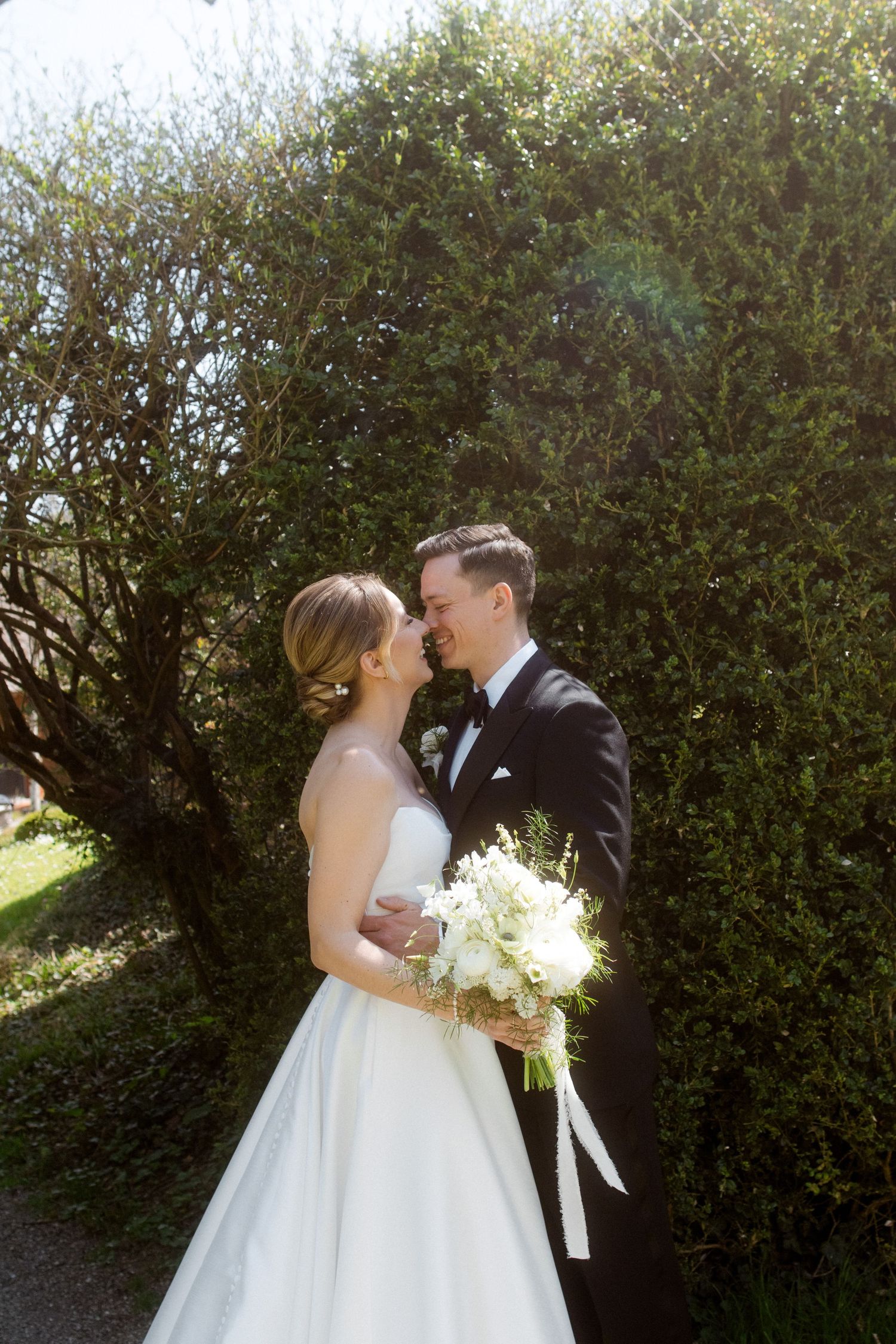 Bride and groom sharing a romantic moment in sunny garden, bride holding white floral bouquet.