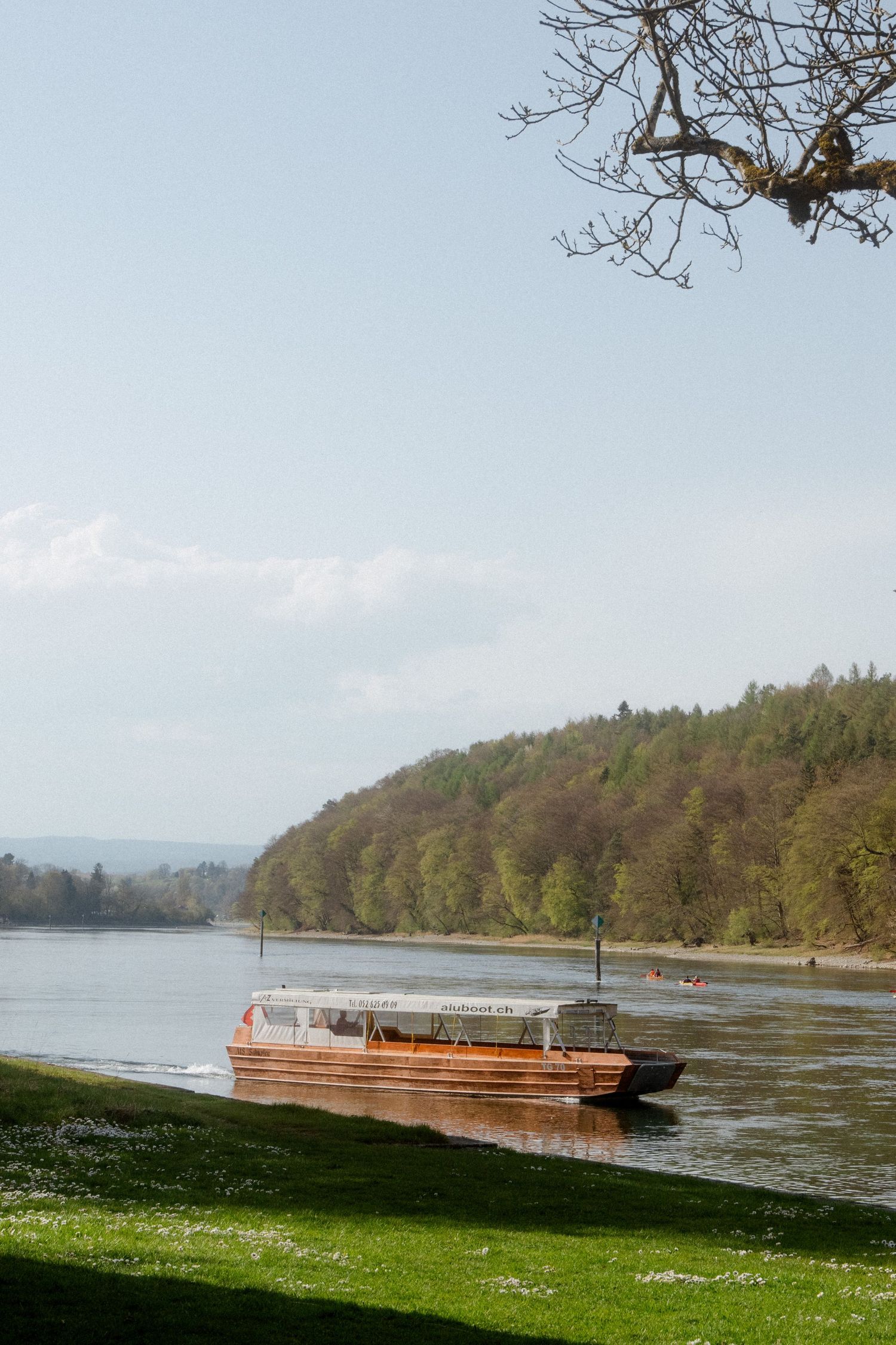 A wooden passenger boat docked on a calm river surrounded by lush forested hills in spring.