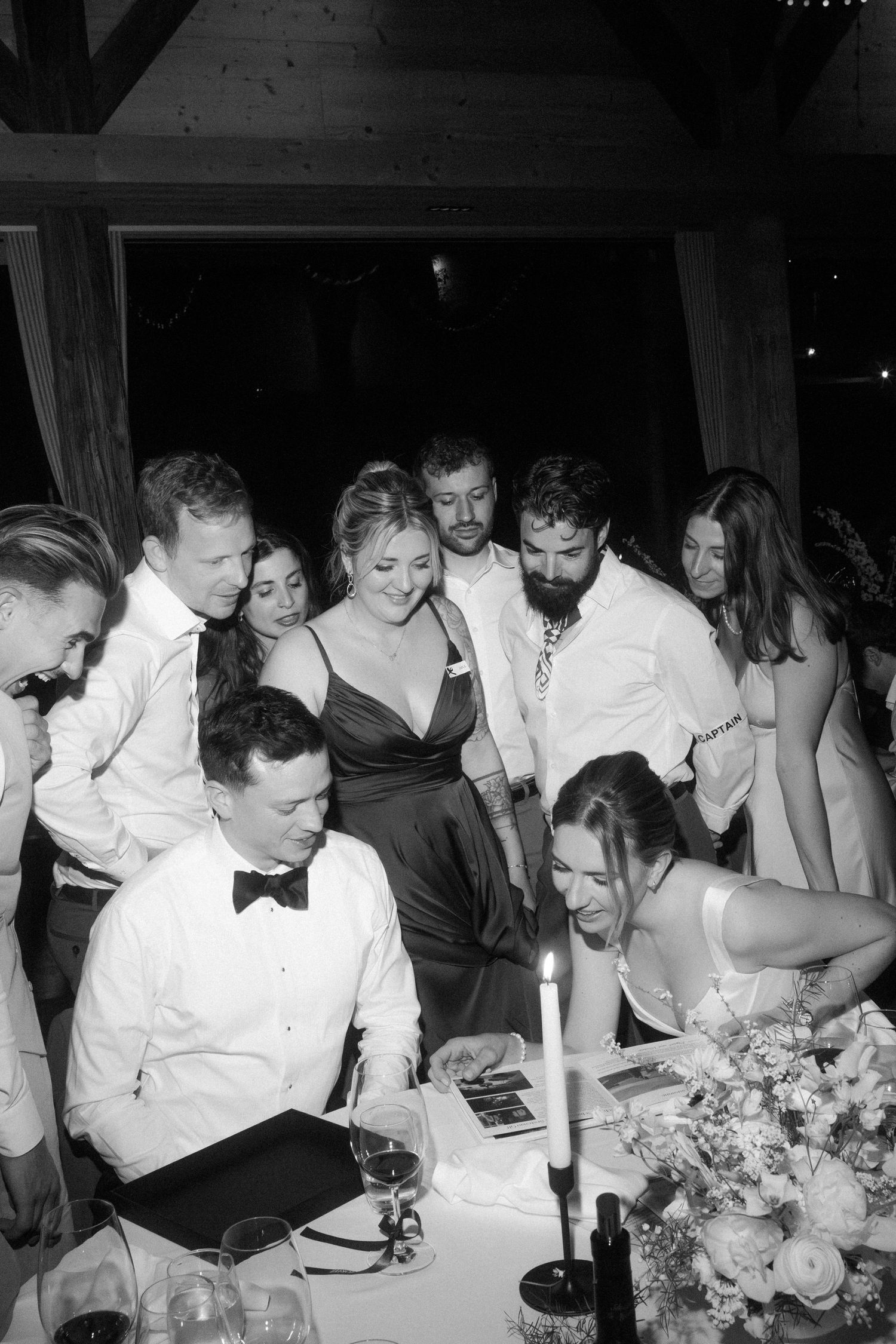 Black and white photo of wedding guests gathered around bride and groom at candlelit reception table.
